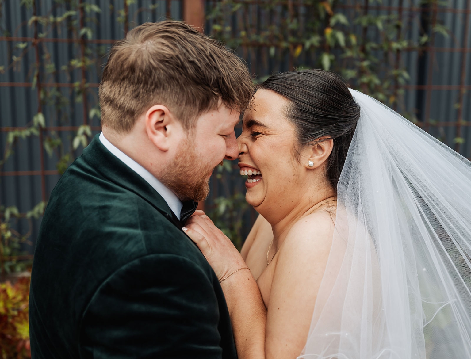 Carley and Corey laughing together with their foreheads touching, the bride wearing a veil and the groom a dark green velvet jacket.
