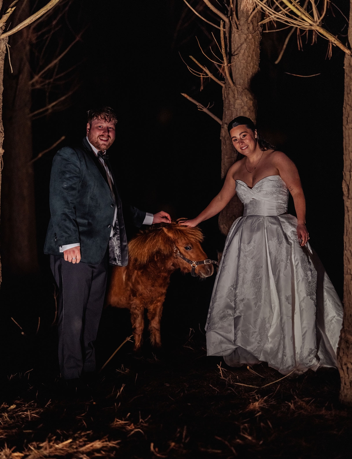 Carley and Corey in wedding attire petting a small pony among trees at night.