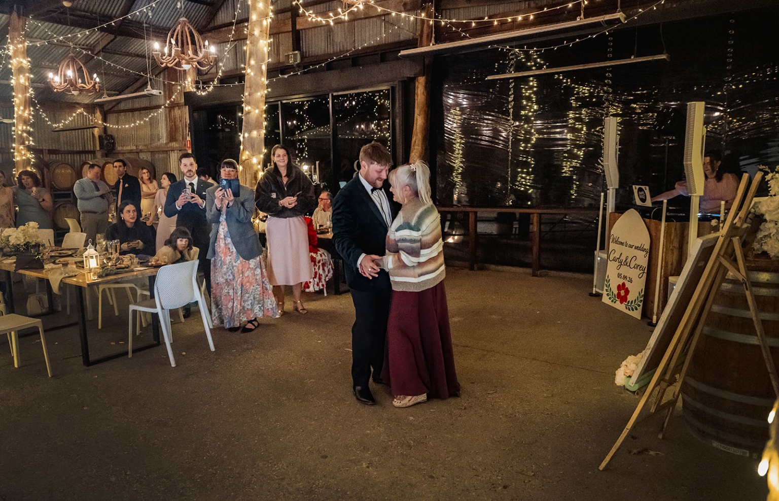 A man dances with a woman at Carley and Corey’s wedding reception while guests watch beneath festoon lights in a rustic barn.