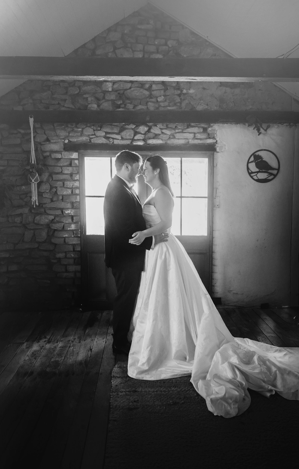 Carley and Corey embracing in front of a backlit door inside a rustic stone cottage, with the bride’s long gown train spread across the timber floor.