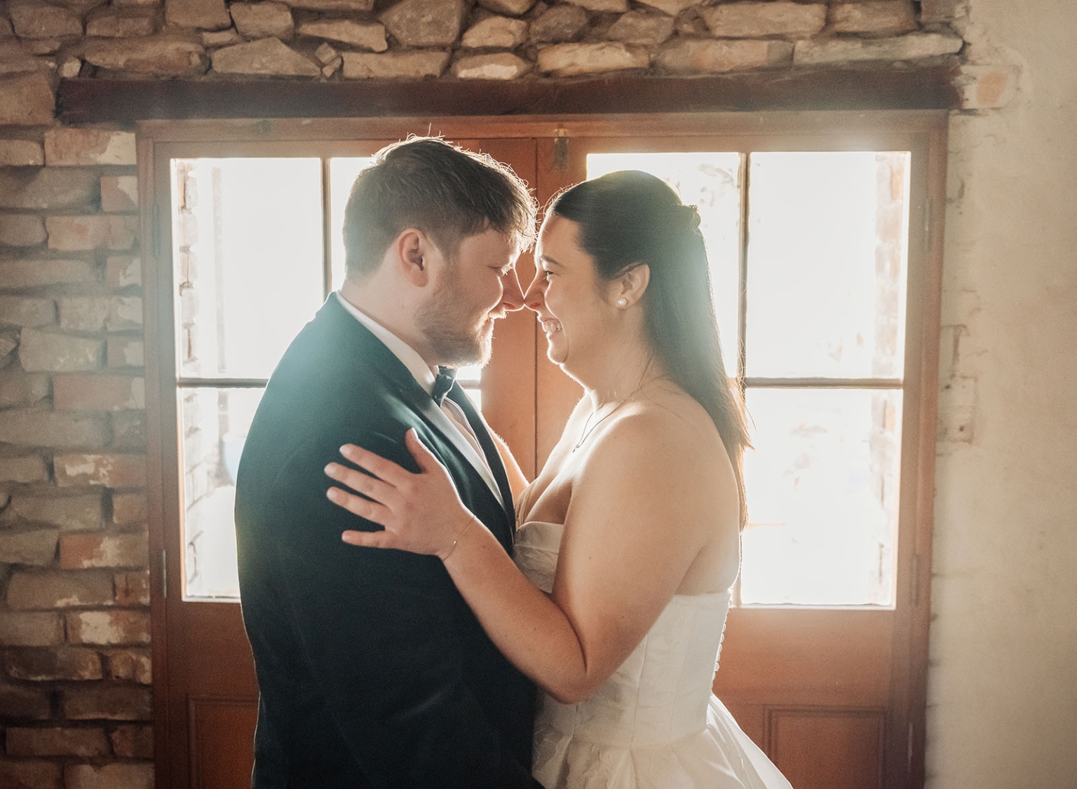Carley and Corey smiling together in front of a sunlit timber-framed window, with Carley in her wedding dress and Corey in a dark suit.