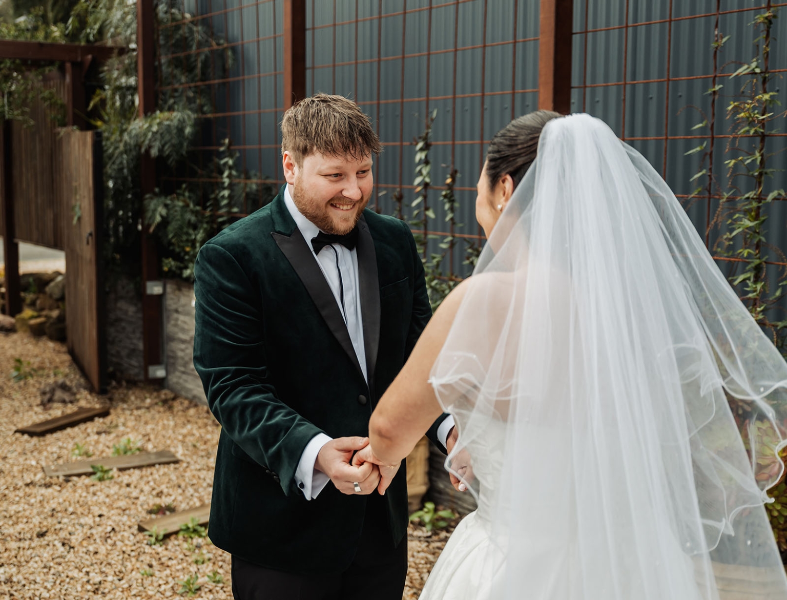 Groom in a dark green velvet tuxedo smiling as he holds hands with the veiled bride during their wedding first look