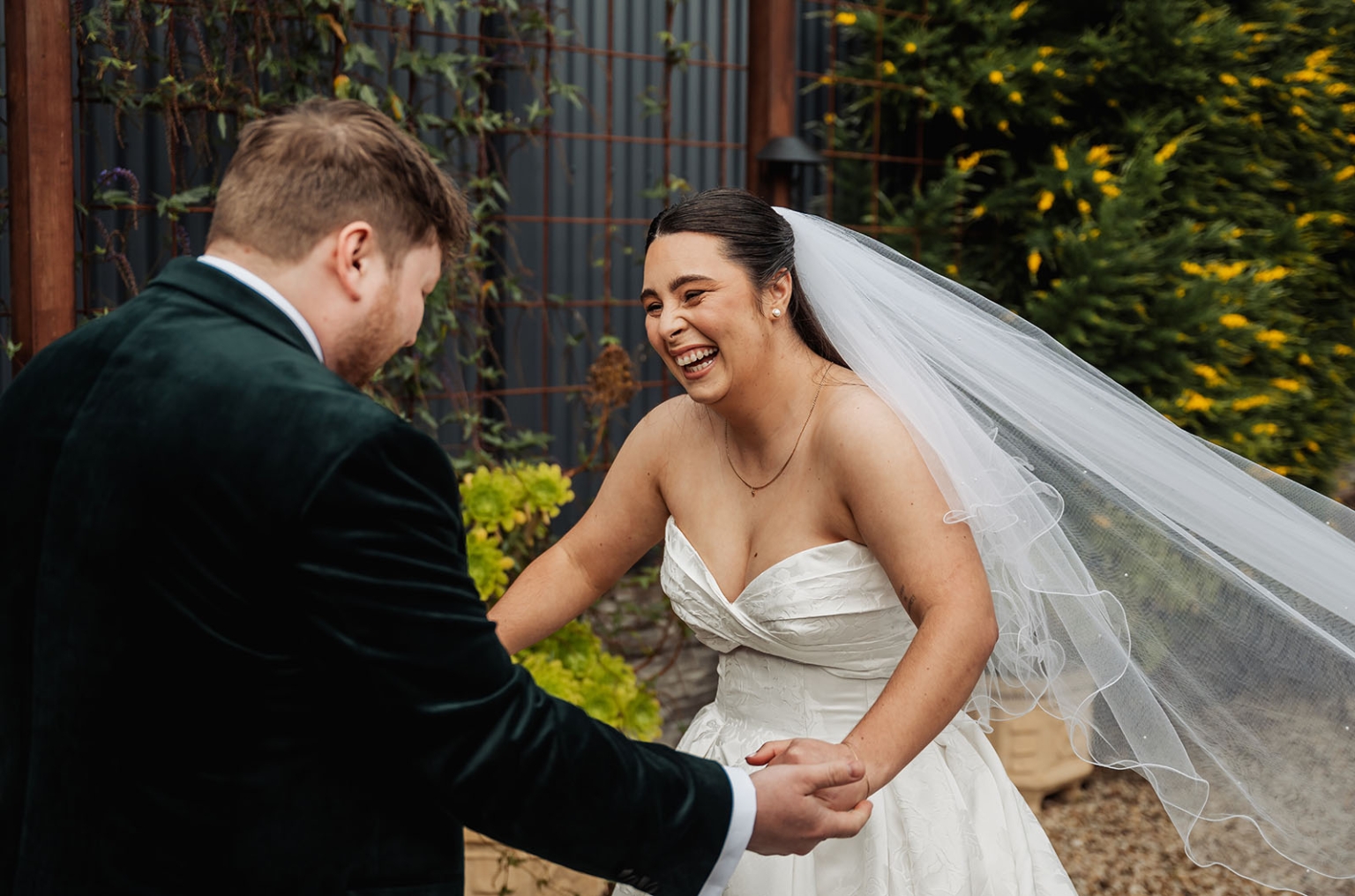 Bride in a white wedding dress and veil laughing while holding hands with the groom in a garden