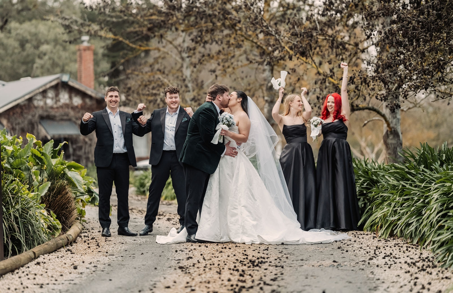Carley and Corey kiss on a garden path as their wedding party celebrates behind them at Barristers Block.