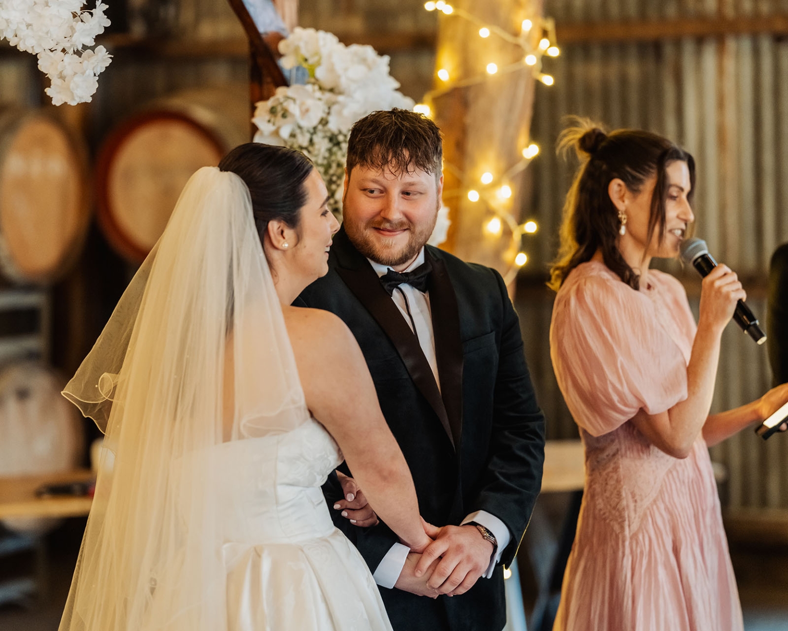 Carley and Corey holding hands during a wedding reception speech, with a woman speaking into a microphone beside them