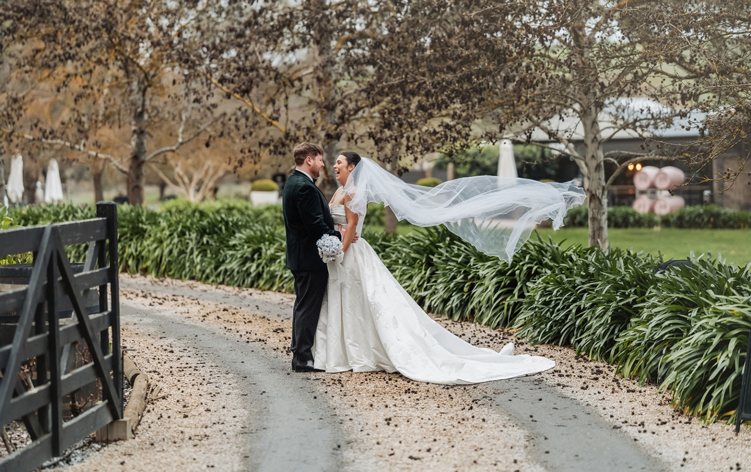 Carley and Corey standing together on a garden path as her long veil blows in the wind at Barristers Block