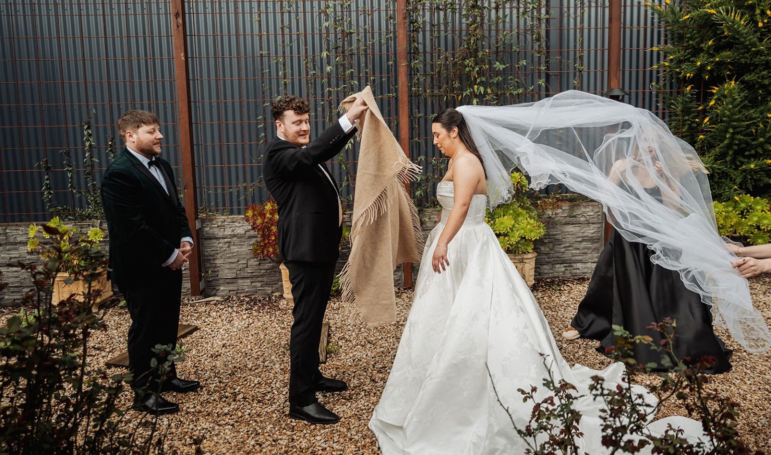 Bride in a strapless white wedding gown with her veil blowing in the wind as two men hold a shawl beside her