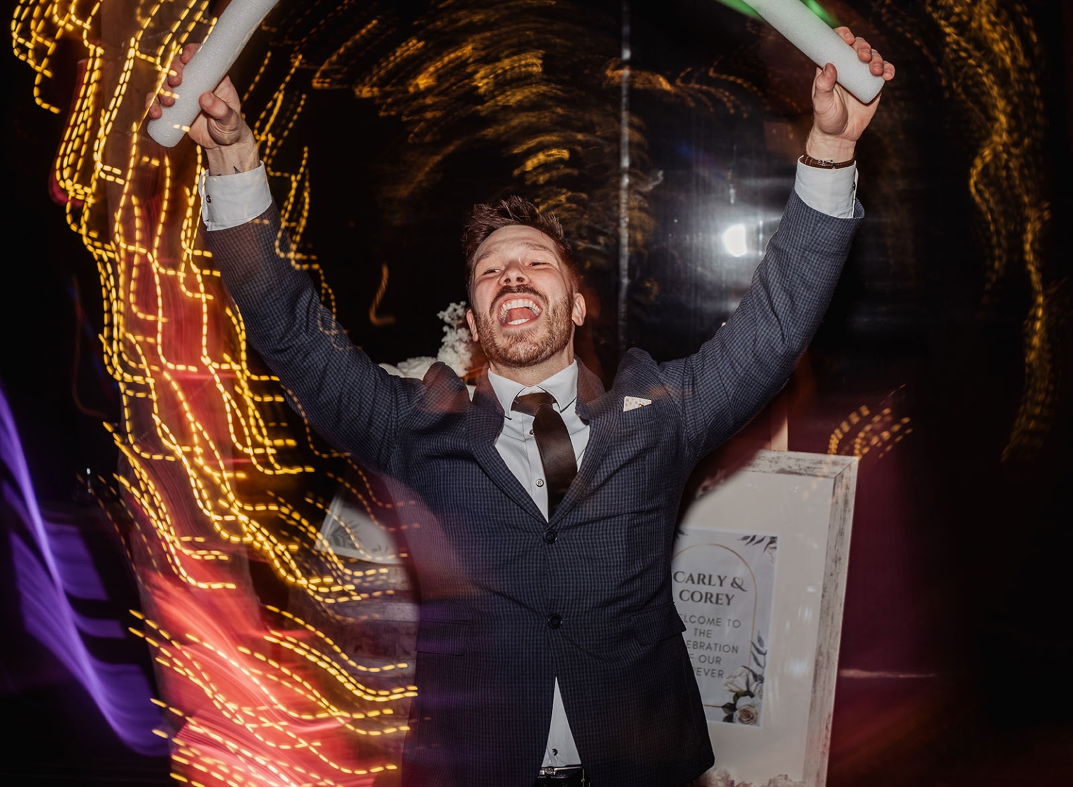 Corey in a suit cheering with illuminated foam sticks amid colourful light trails at the wedding reception.