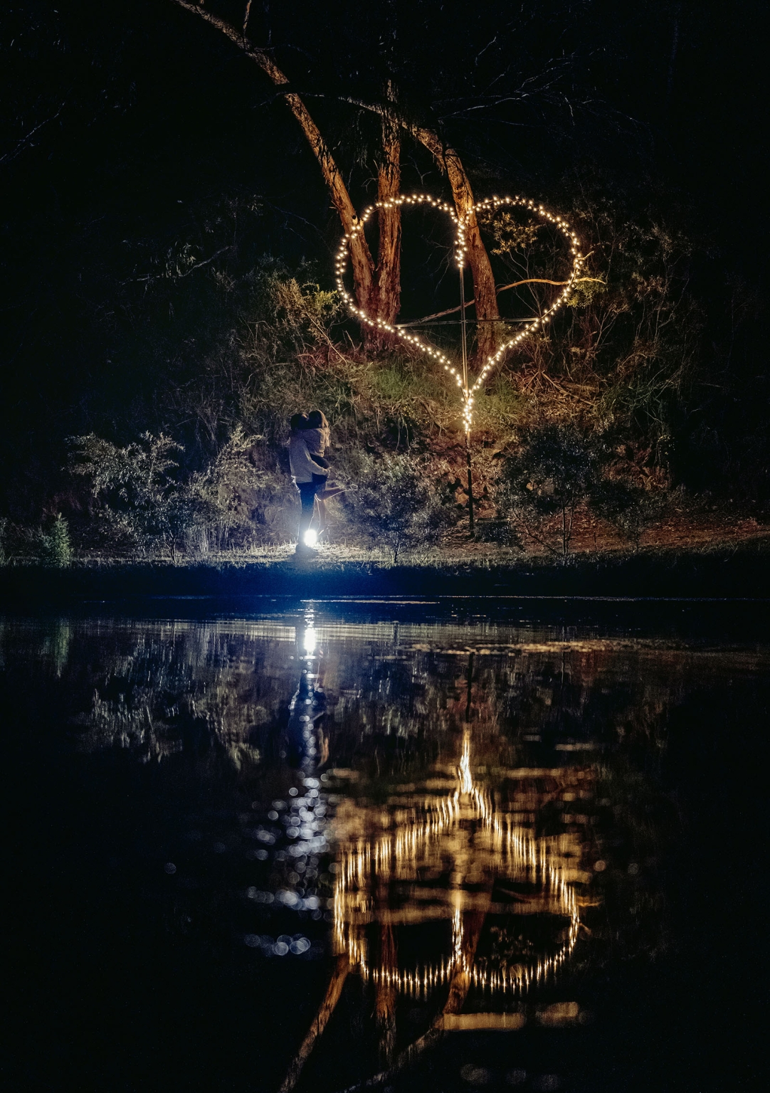 tiny house on the prairie proposal - kyle & bianca 17 Couple embracing beside a large heart-shaped fairy light display reflected in a pond at night