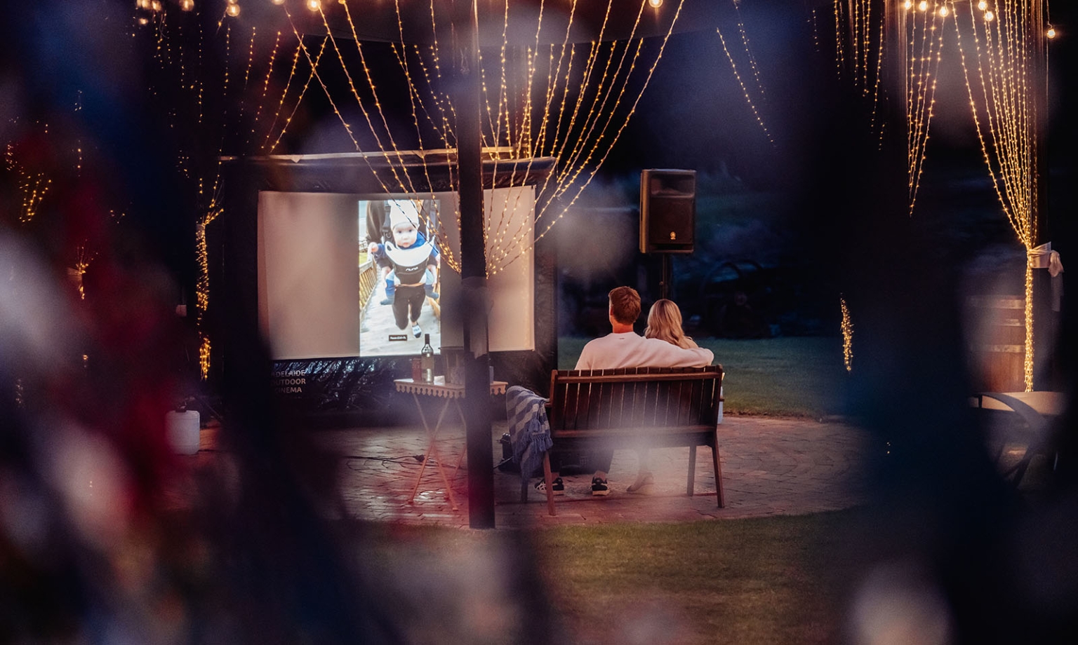 tiny house on the prairie proposal - kyle & bianca 3 A couple sit together on a bench watching a projected relationship video beneath fairy lights at Tiny House on the Prairie.