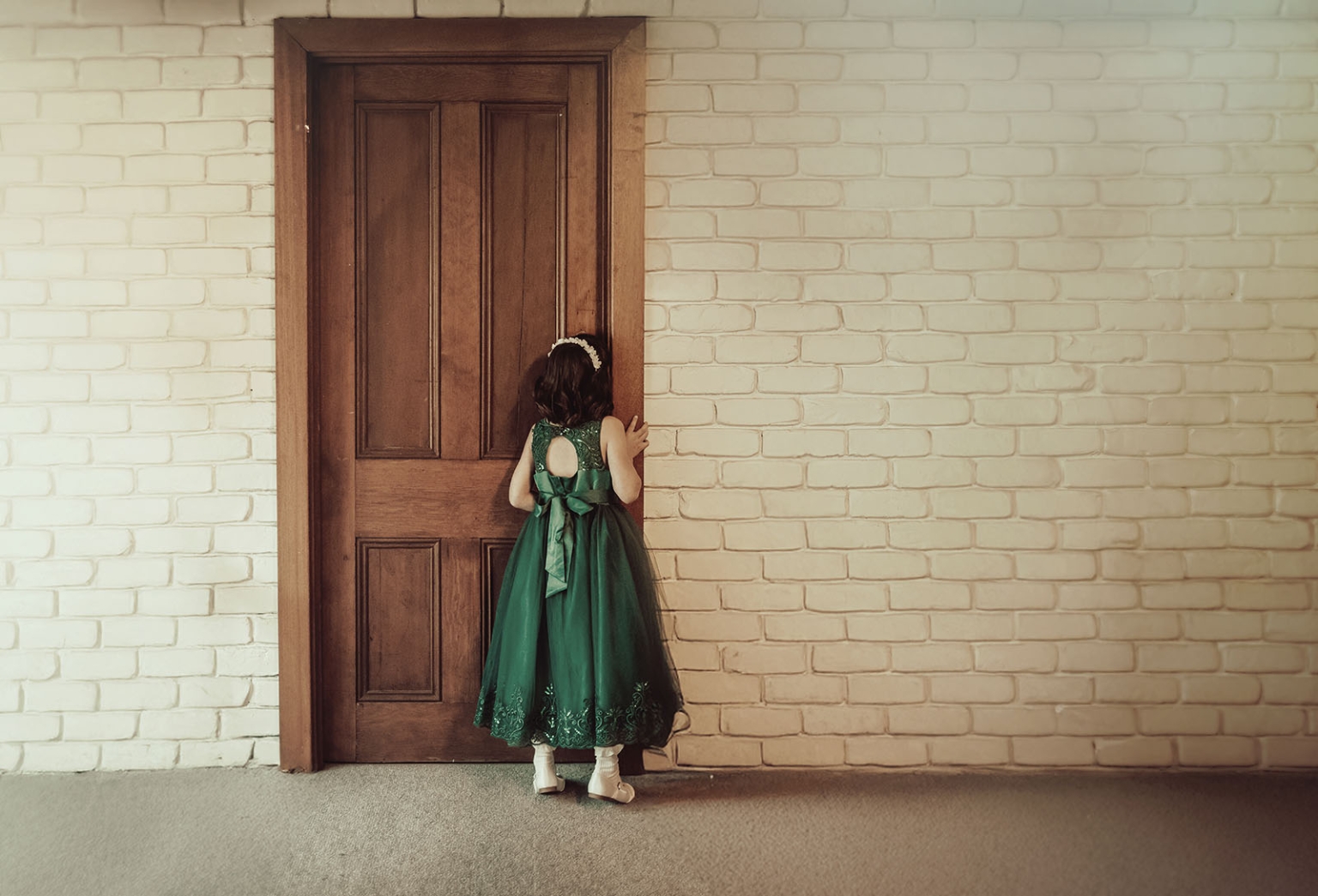 Flower girl in a green dress standing at a timber door against a white painted brick wall