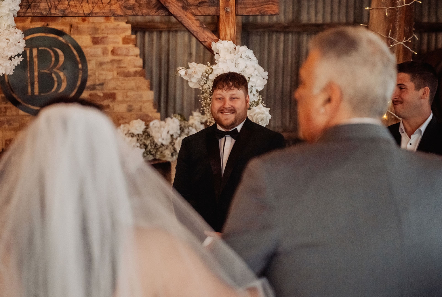 Groom smiling at the altar as the bride, accompanied by an older man, approaches through white floral arrangements at Barristers Block.