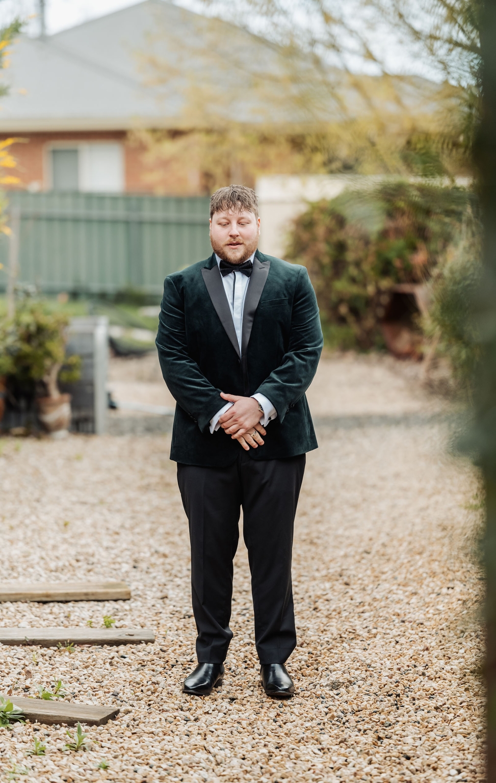 Groom in a dark green velvet tuxedo jacket and bow tie standing on a gravel garden path
