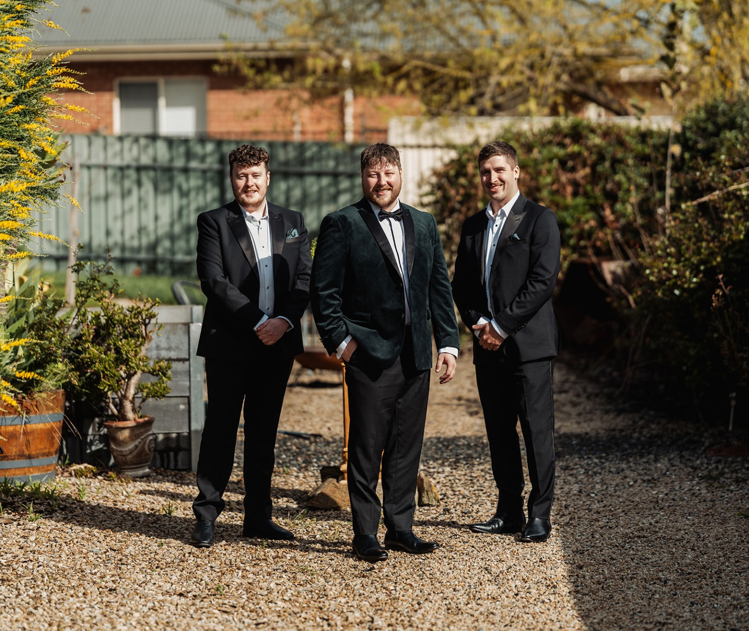 Groom in a dark green velvet dinner jacket standing with two groomsmen in a cottage garden
