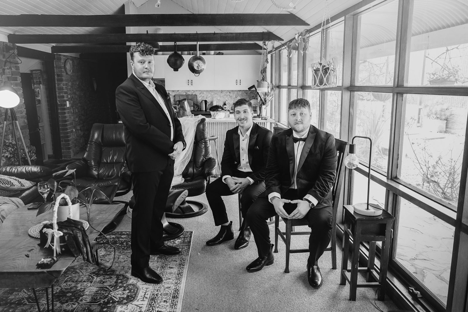 Three men in formal suits seated and standing in a rustic cottage living room during wedding preparations