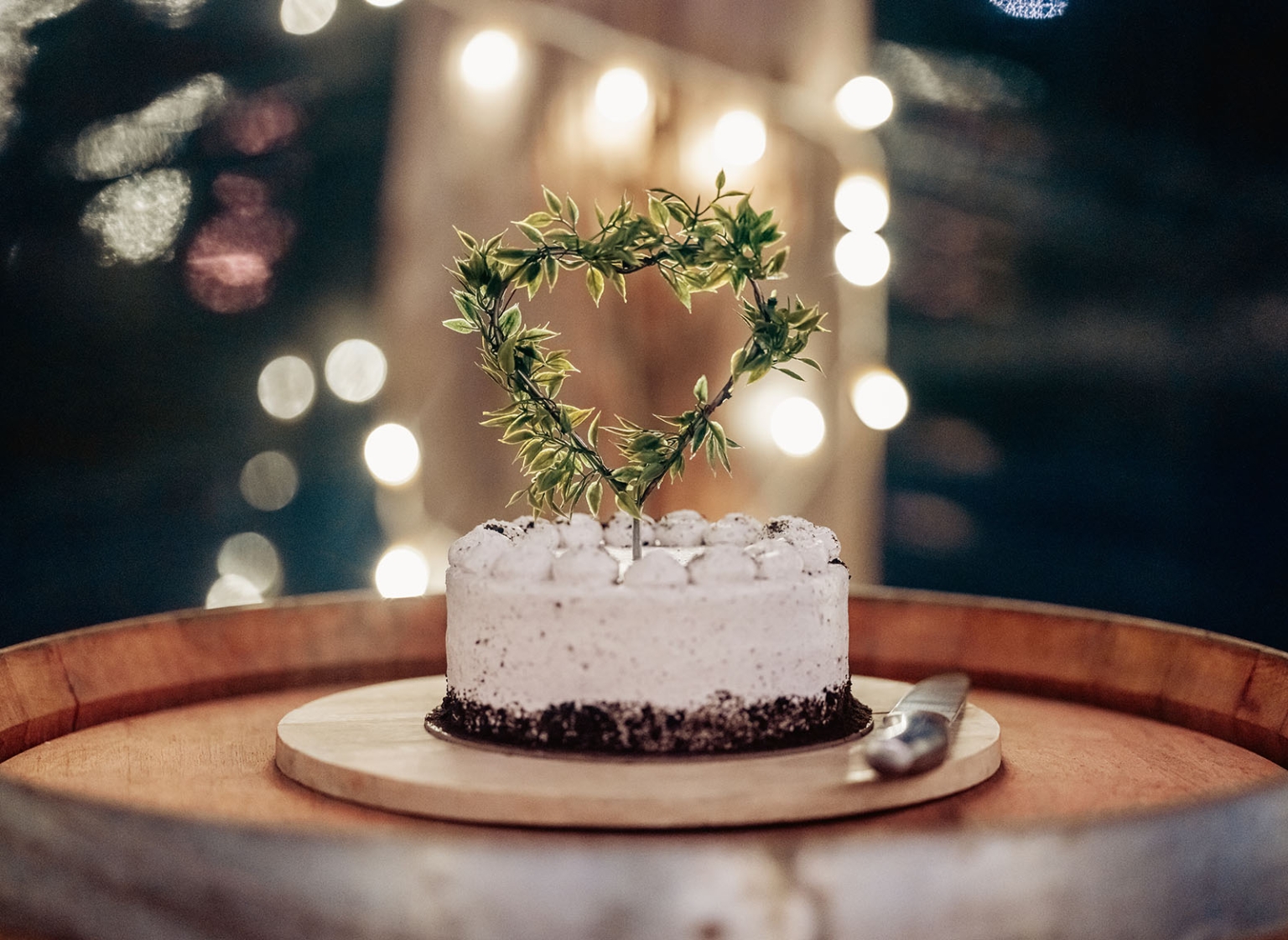 Small white wedding cake with a leafy heart topper on a wooden barrel, lit by warm festoon lights.