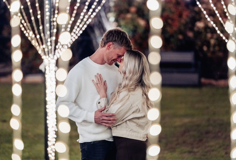 Kyle and Bianca embracing beneath strings of fairy lights, with Bianca’s engagement ring visible.