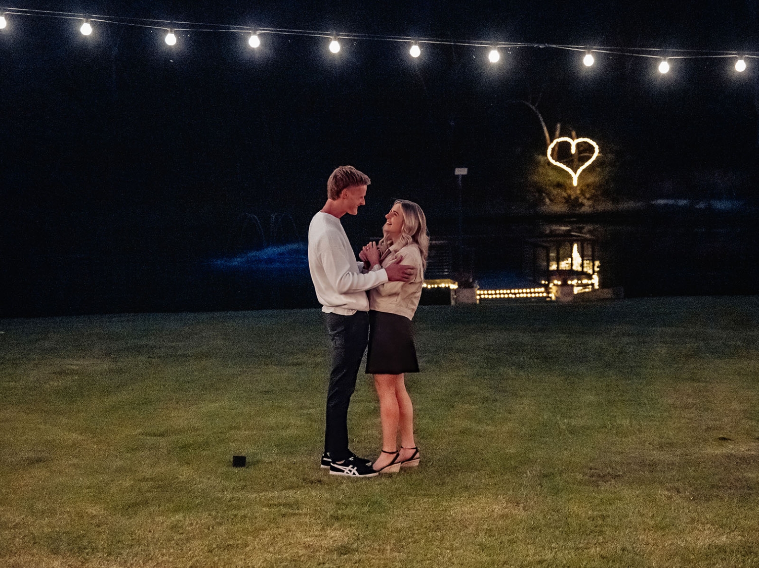 tiny house on the prairie proposal - kyle & bianca 7 Kyle and Bianca embracing on a lawn beneath fairy lights and a glowing heart display at Tiny House on the Prairie