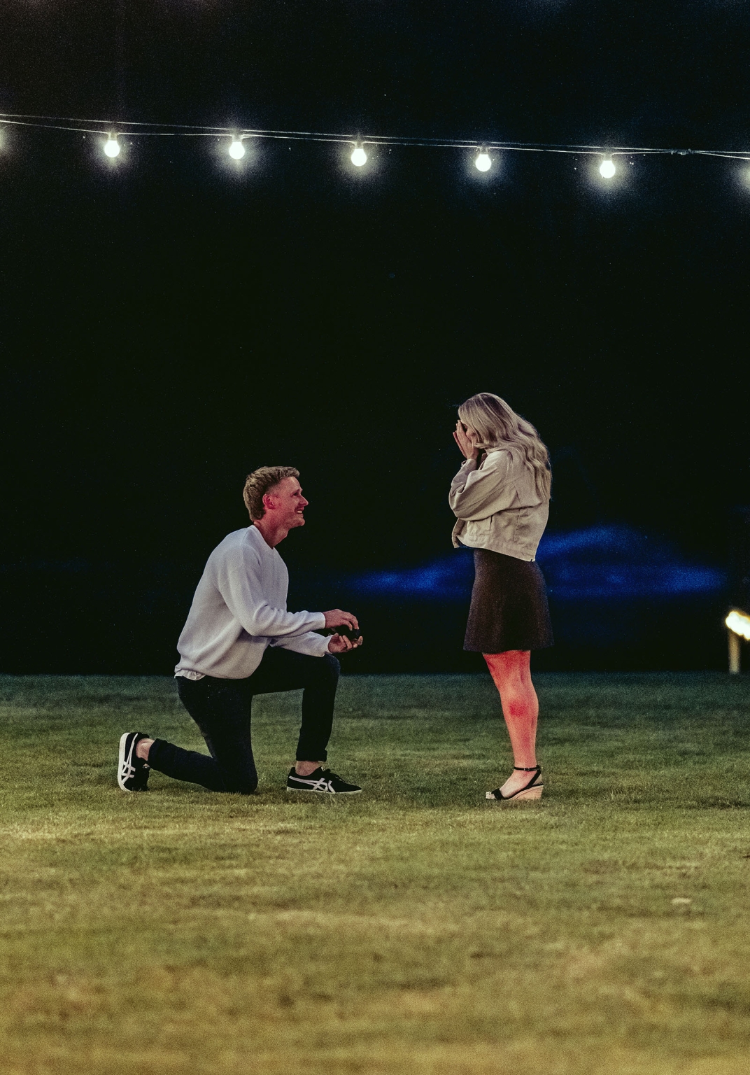 tiny house on the prairie proposal - kyle & bianca 5 Kyle kneels to propose to Bianca on a lawn beneath glowing string lights at night.