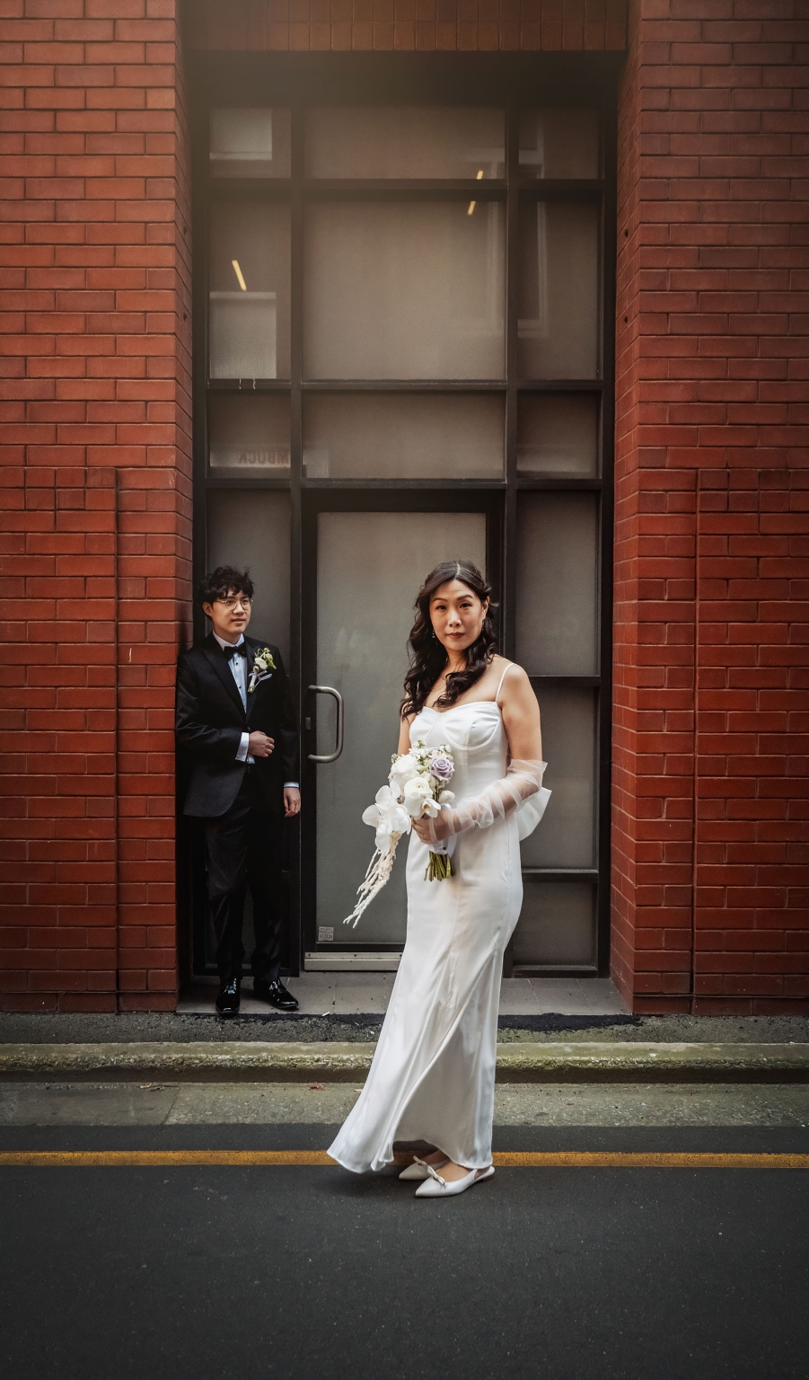 marriage rego office wedding – teri & thomas 19 Bride holding a white and lavender bouquet stands in front of a brick building while the groom waits in the doorway behind her.