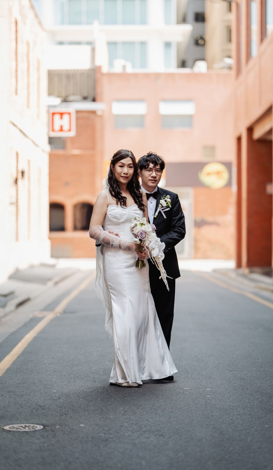 marriage rego office wedding – teri & thomas 18 Teri and Thomas standing together in a city laneway, with Teri holding a pastel wedding bouquet in her white gown and Thomas wearing a black tuxedo.