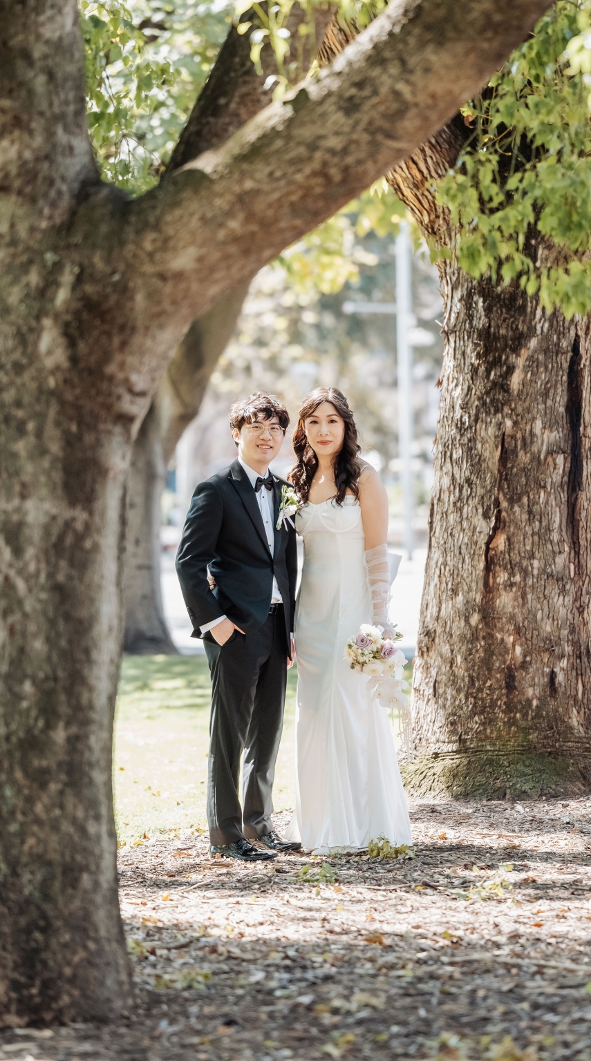 marriage rego office wedding – teri & thomas 21 Teri and Thomas standing together between large tree trunks, with Teri holding a pastel wedding bouquet.