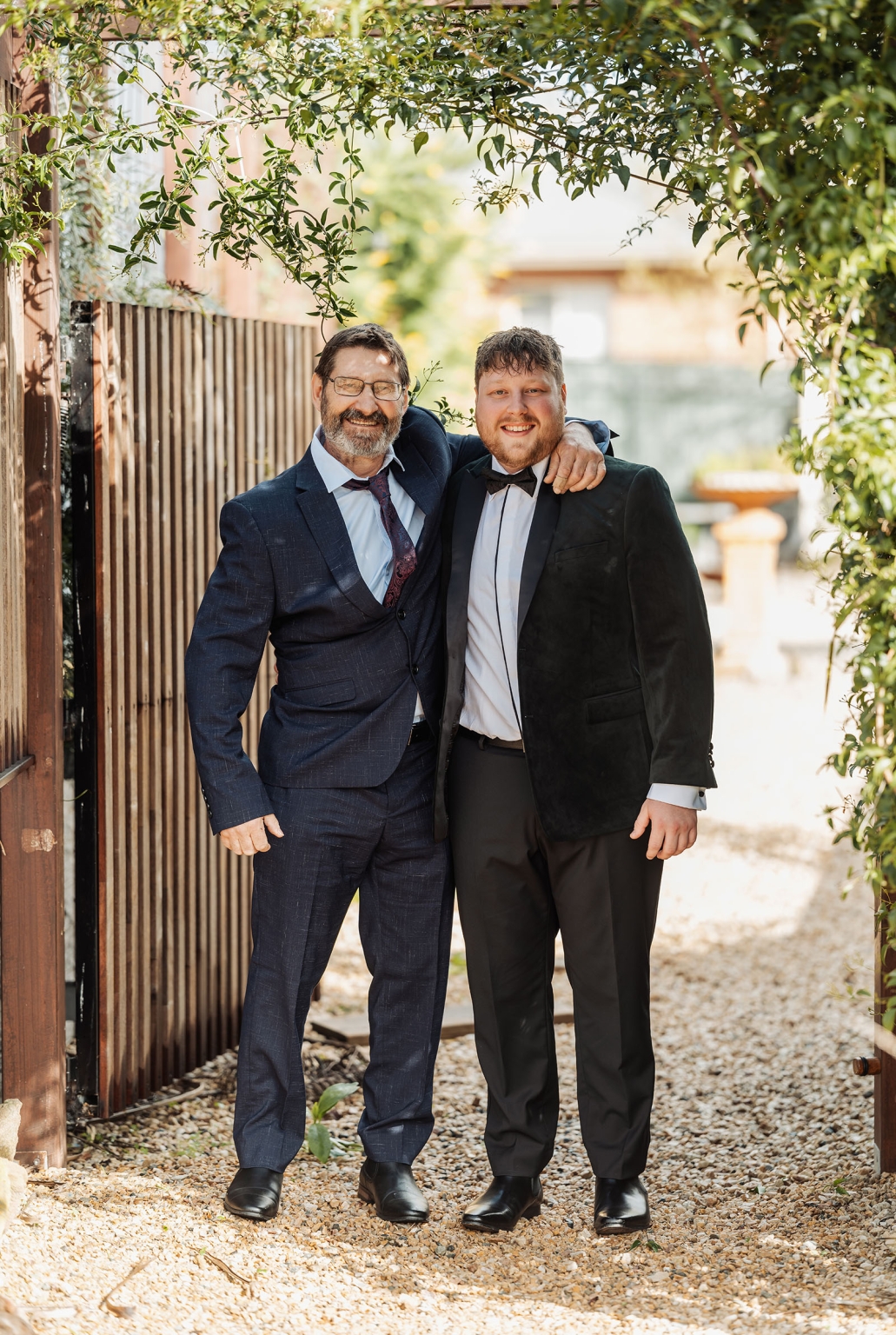 Two men in formal wedding attire smiling with their arms around each other beneath leafy branches