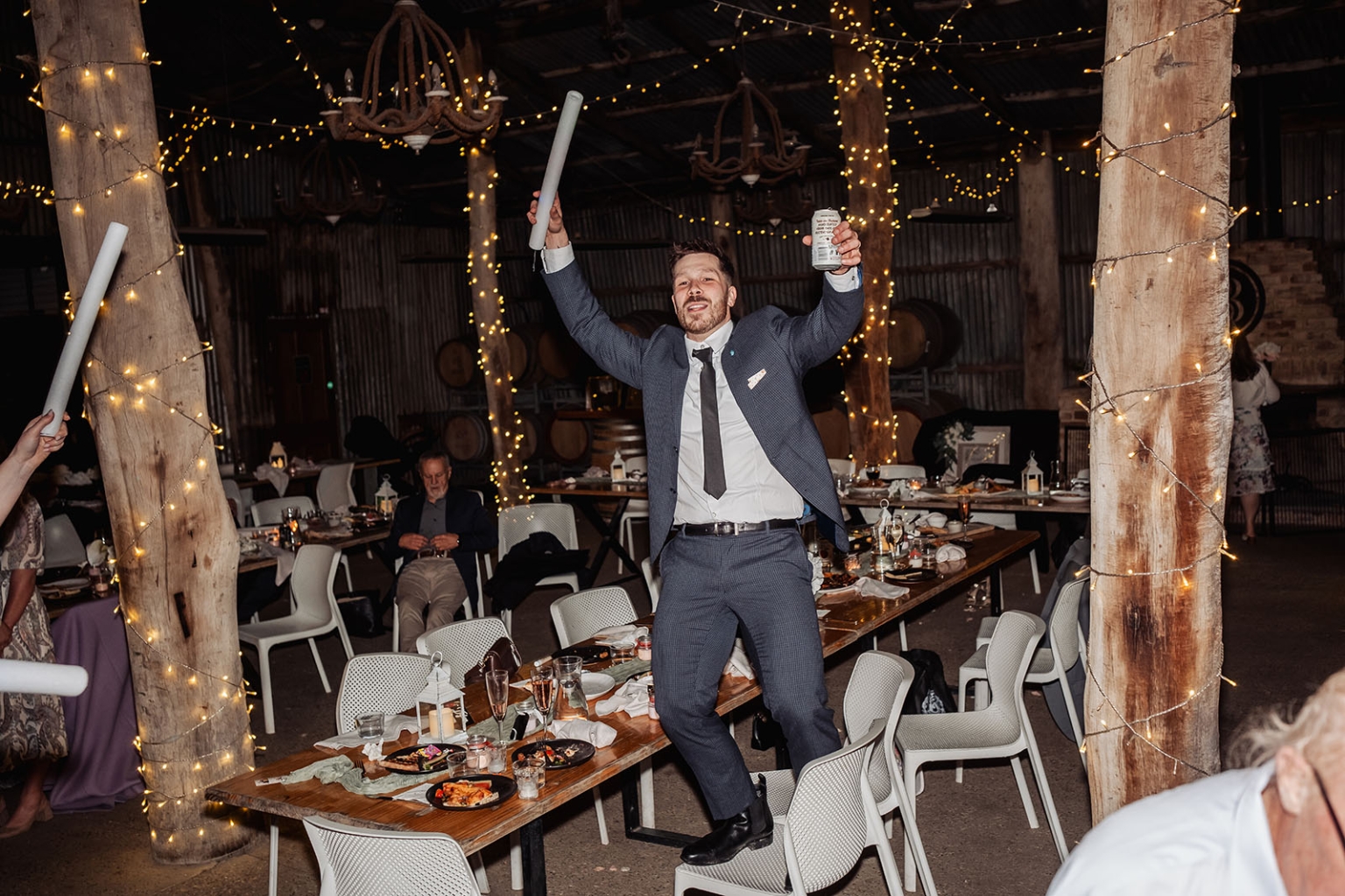 Wedding guest in a blue suit dancing between reception tables with a foam baton and drink can under fairy lights