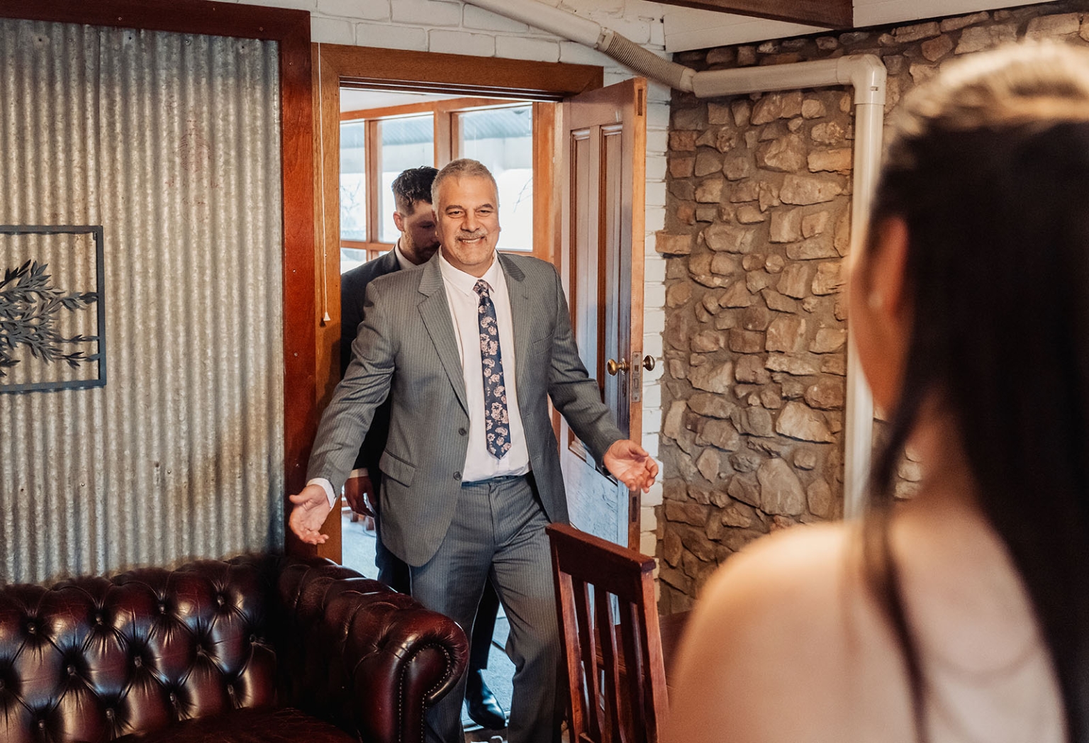 Smiling man in a grey suit entering a rustic stone-walled cottage room during wedding preparations