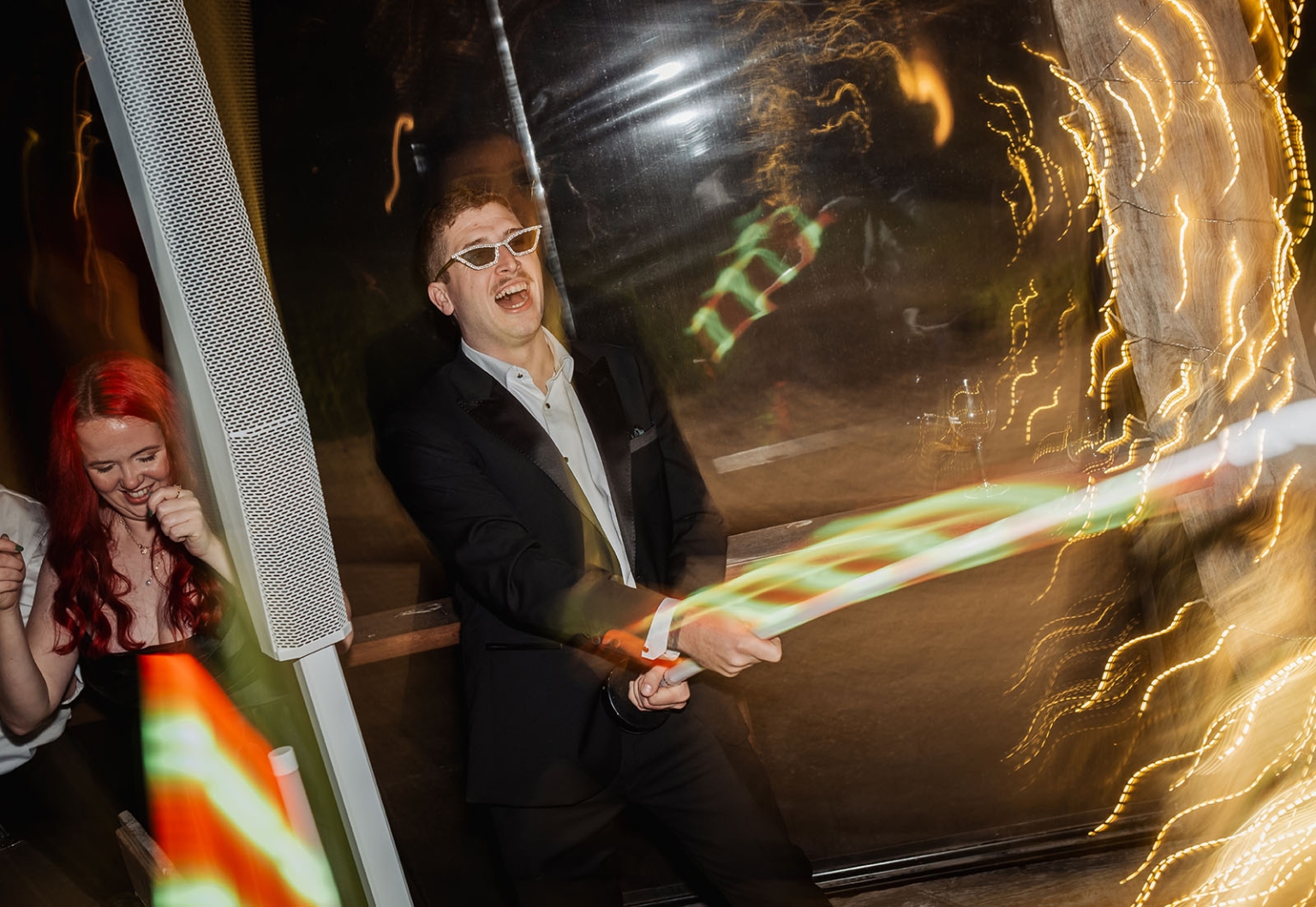 Man in a black suit and novelty glasses laughing beneath colourful light trails at a wedding party
