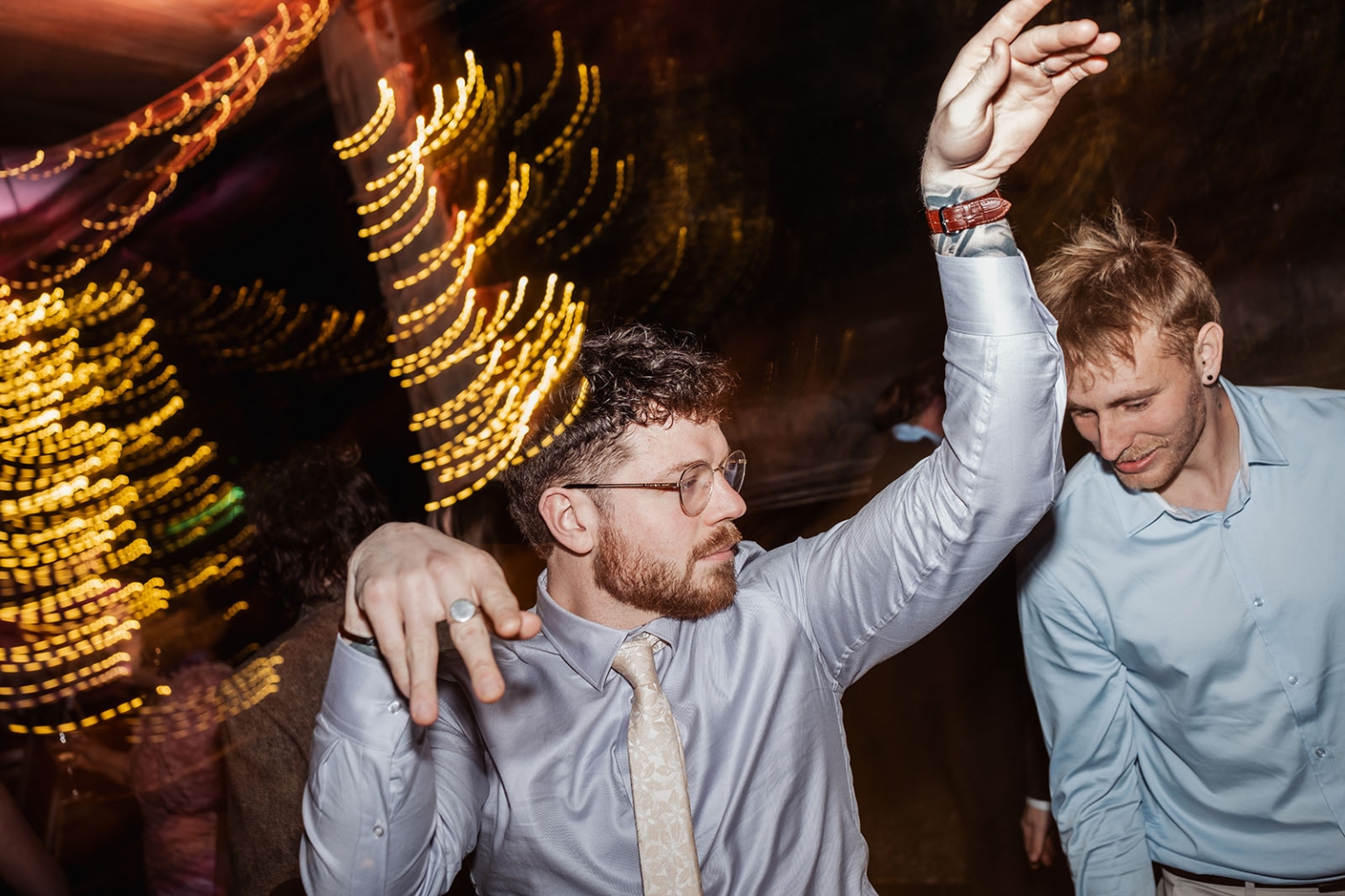 Two male wedding guests dancing beneath warm fairy lights at the reception