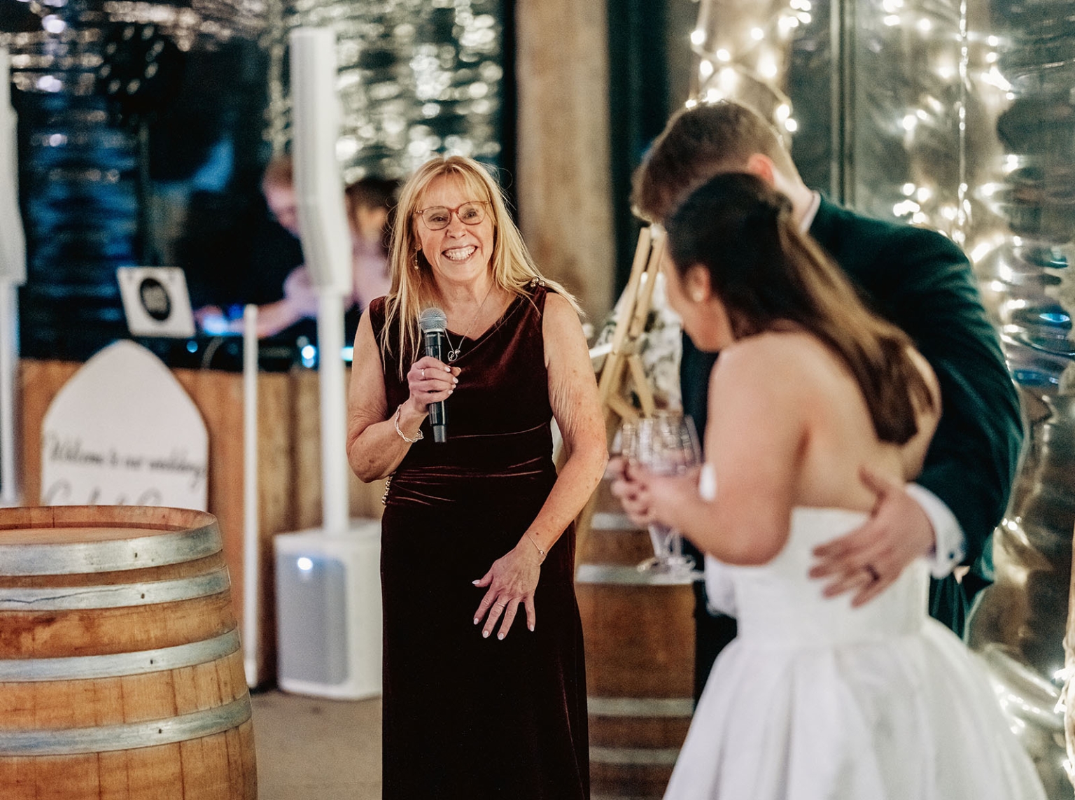 Woman giving a wedding reception speech as the bride and groom embrace beside fairy lights