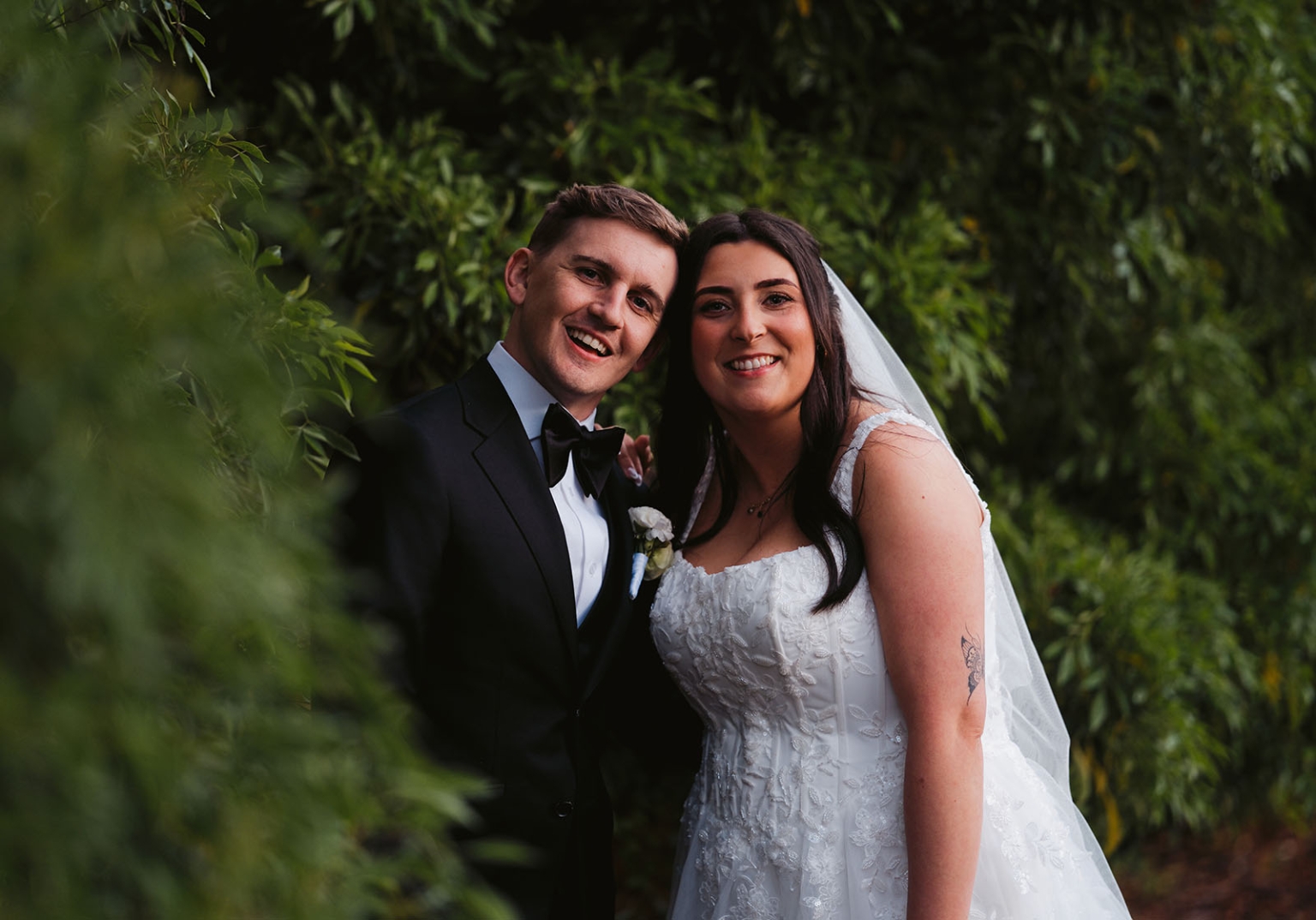 beach road winery wedding – alysha & nik 43 Alysha and Nik smiling together among lush green foliage, with Alysha in a lace wedding dress and veil and Nik in a black tuxedo.