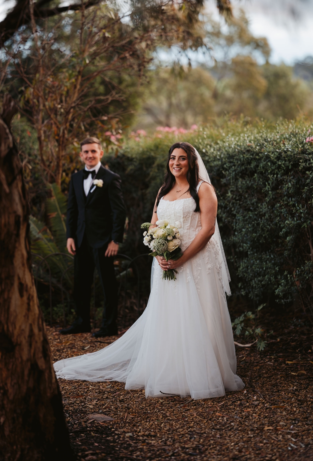 beach road winery wedding – alysha & nik 31 Bride Alysha in a white wedding dress holding a bouquet, with groom Nik standing behind her in a leafy garden at Beach Road Winery