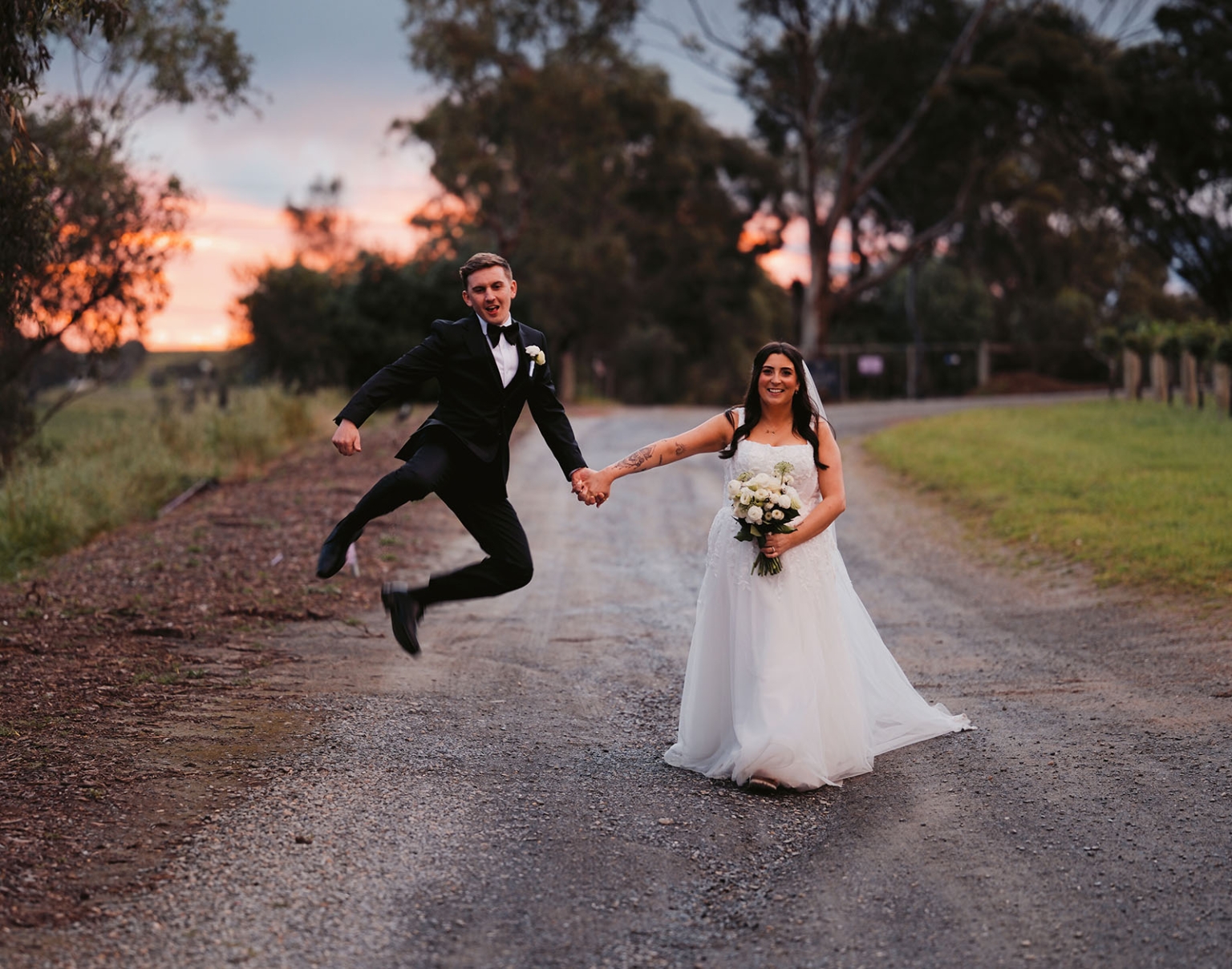 beach road winery wedding – alysha & nik 40 Nik jumps while holding Alysha’s hand on a tree-lined gravel road at sunset, as she holds her white wedding bouquet.