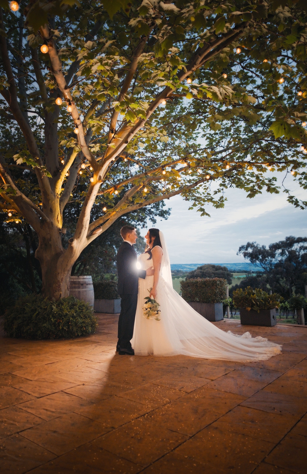 beach road winery wedding – alysha & nik 46 Alysha and Nik stand beneath a large tree strung with warm fairy lights at Beach Road Winery, with her long wedding dress train spread across the terrace.