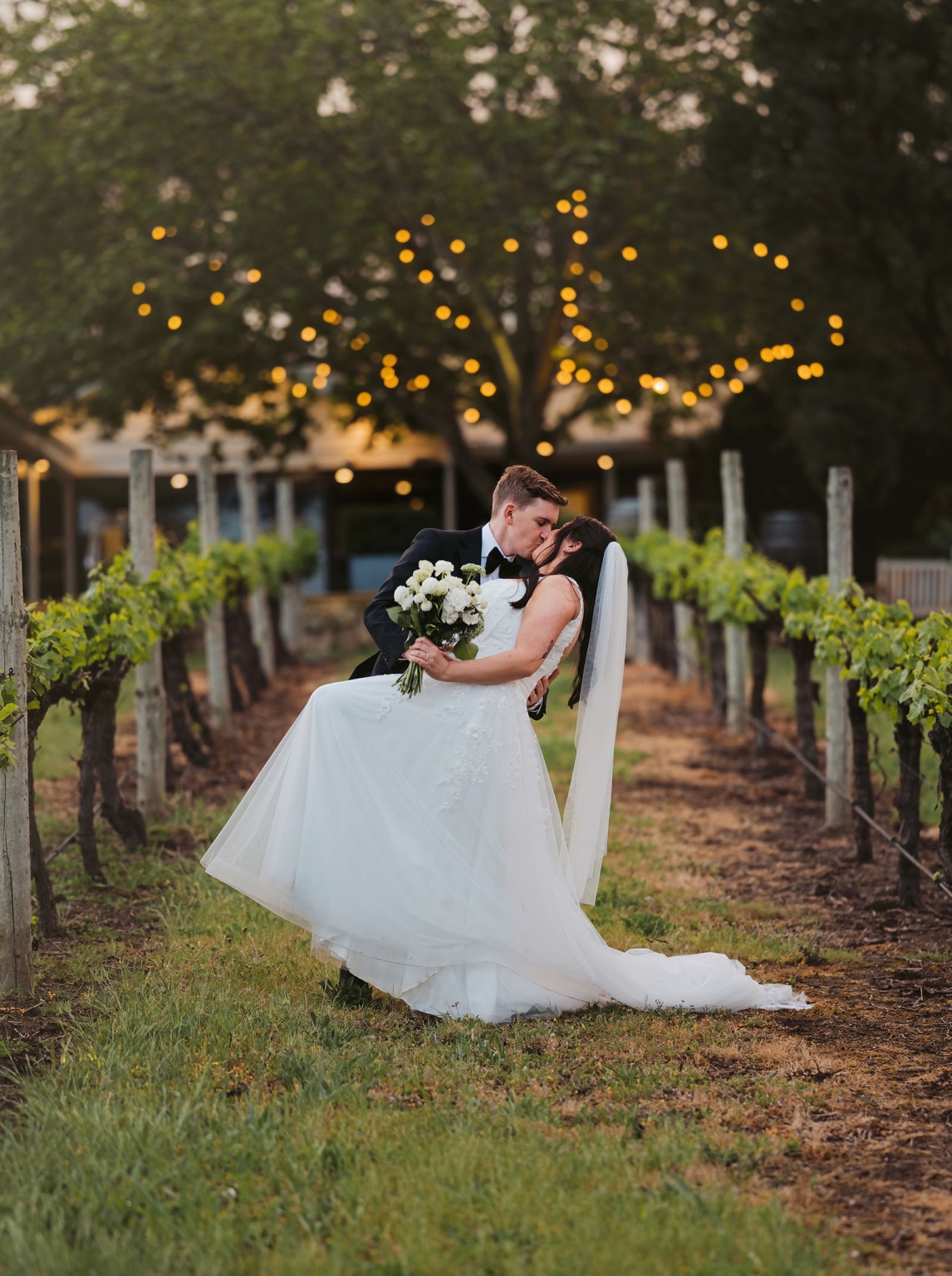 beach road winery wedding – alysha & nik 35 Alysha and Nik kissing between vineyard rows, with Alysha holding a white bouquet beneath glowing festoon lights at Beach Road Winery.