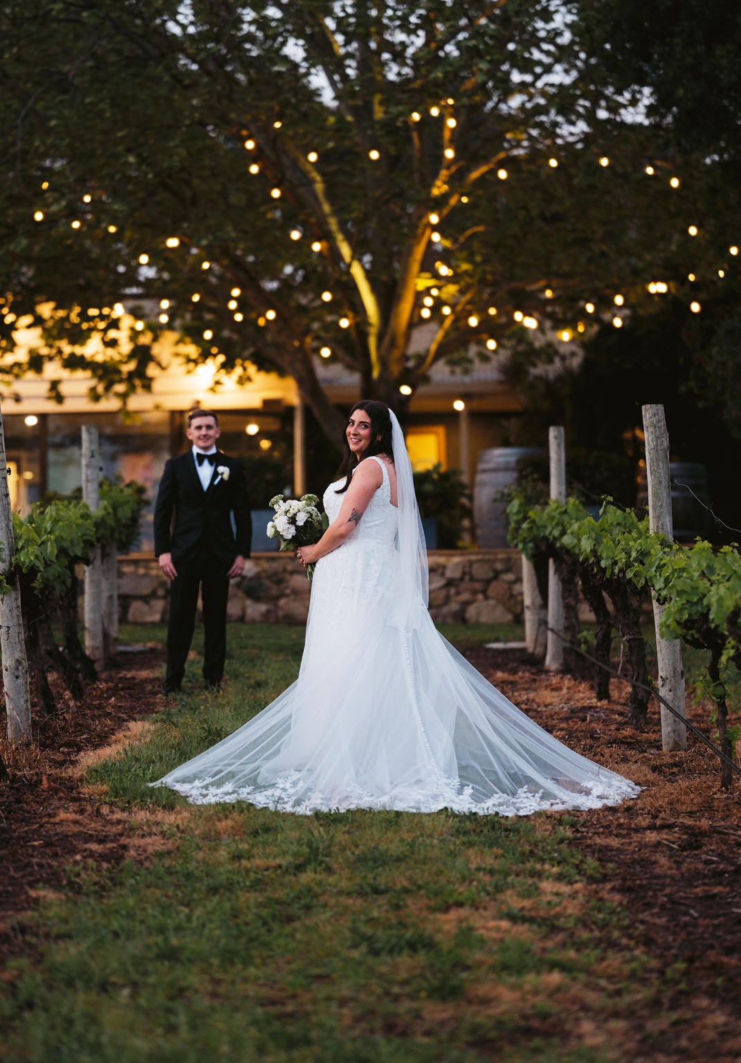 beach road winery wedding – alysha & nik 45 Bride Alysha in a white gown and long veil holds a bouquet between vineyard rows, with groom Nik beneath festoon lights behind her at Beach Road Winery.