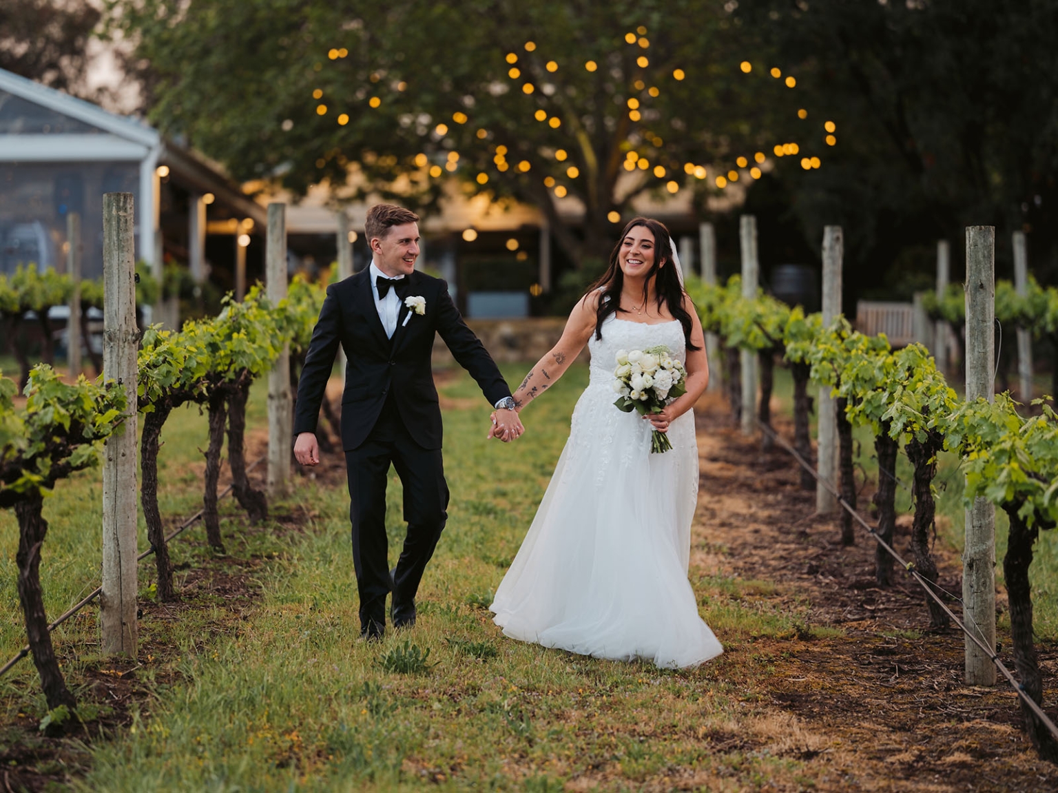 beach road winery wedding – alysha & nik 34 Alysha and Nik walk hand in hand between vineyard rows, with Alysha holding a white bouquet in her wedding dress.