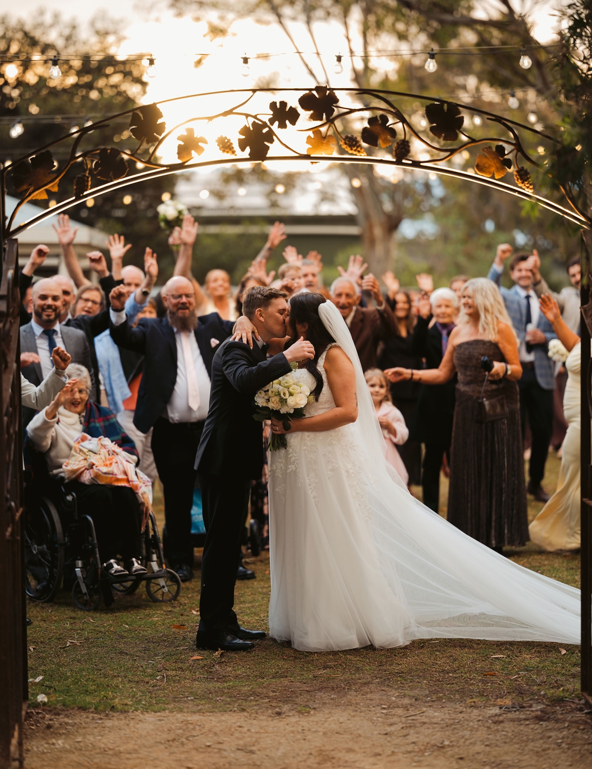 beach road winery wedding – alysha & nik 29 Alysha and Nik kiss beneath a grapevine-decorated arch as wedding guests cheer behind them at Beach Road Winery.