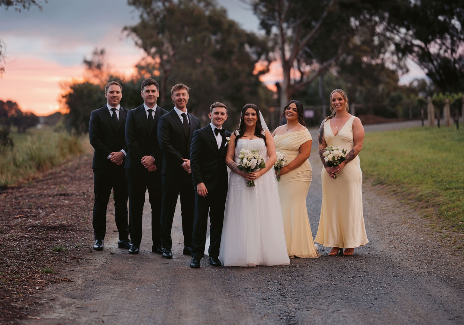 beach road winery wedding – alysha & nik 38 Alysha and Nik stand with their wedding party on a tree-lined gravel path at sunset, with groomsmen in black suits and bridesmaids in pale yellow dresses.