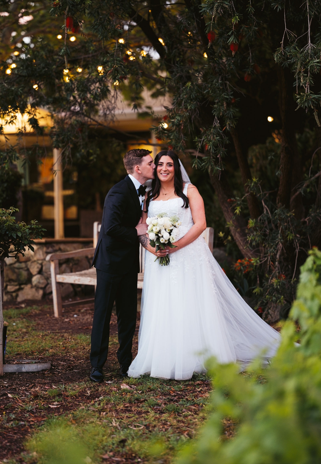 beach road winery wedding – alysha & nik 44 Nik kisses Alysha on the cheek as she holds a white bouquet beneath leafy trees and warm lights at Beach Road Winery.