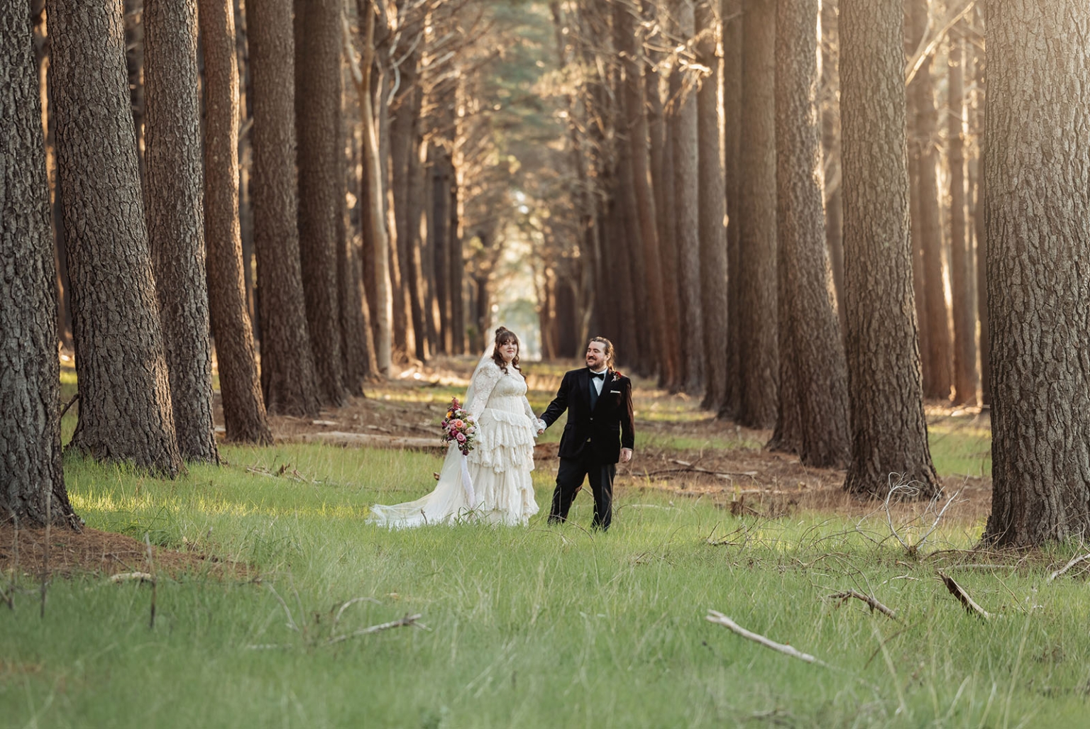 burbrook wedding – amber & angus | kuitpo forest wedding 49 Amber and Angus holding hands among tall pine trees at Burbrook Forest, with Amber in a layered white wedding dress holding a colourful bouquet.