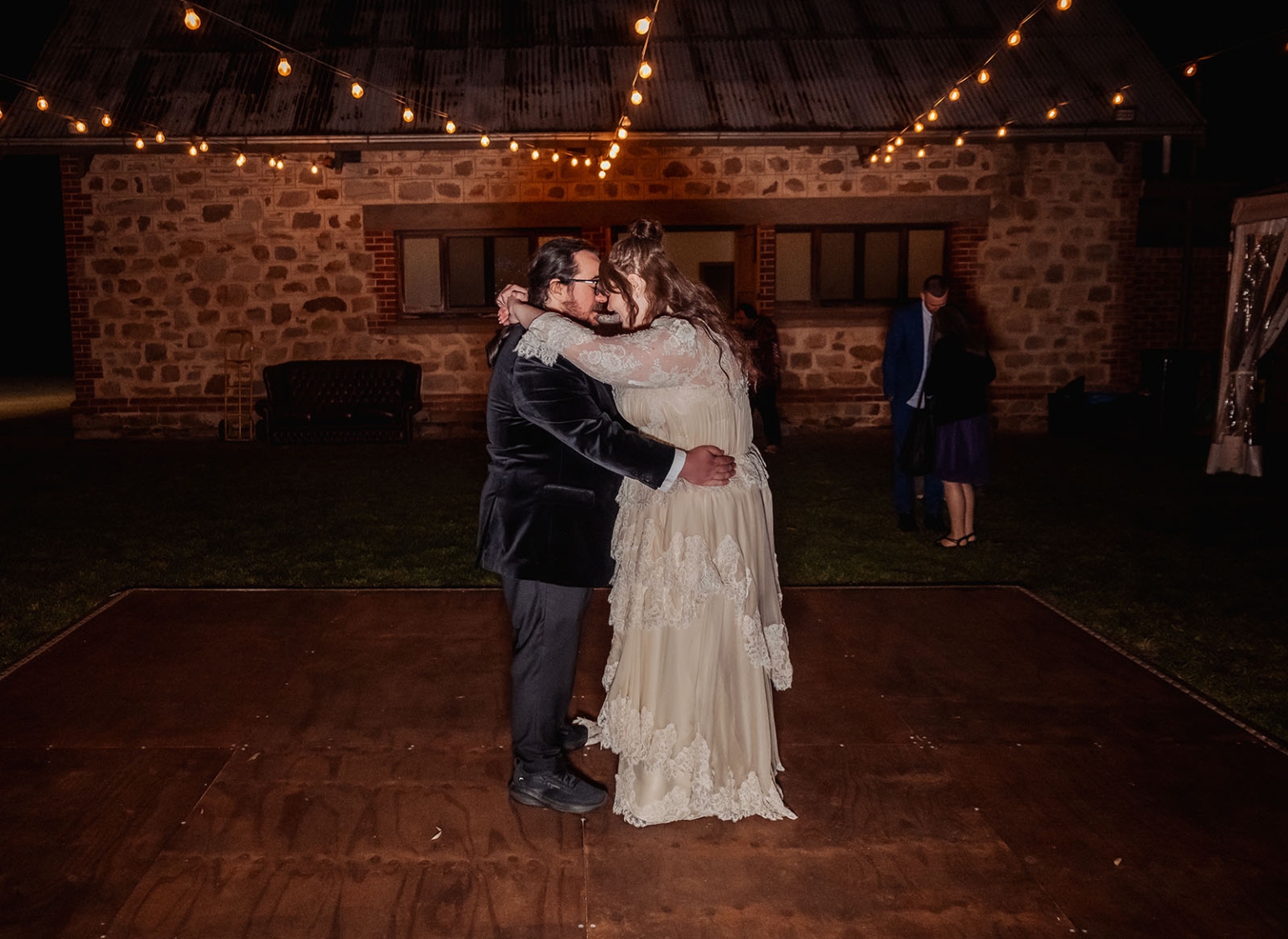 burbrook wedding – amber & angus | kuitpo forest wedding 68 Amber and Angus embracing during their first dance beneath festoon lights outside a stone building at Burbrook