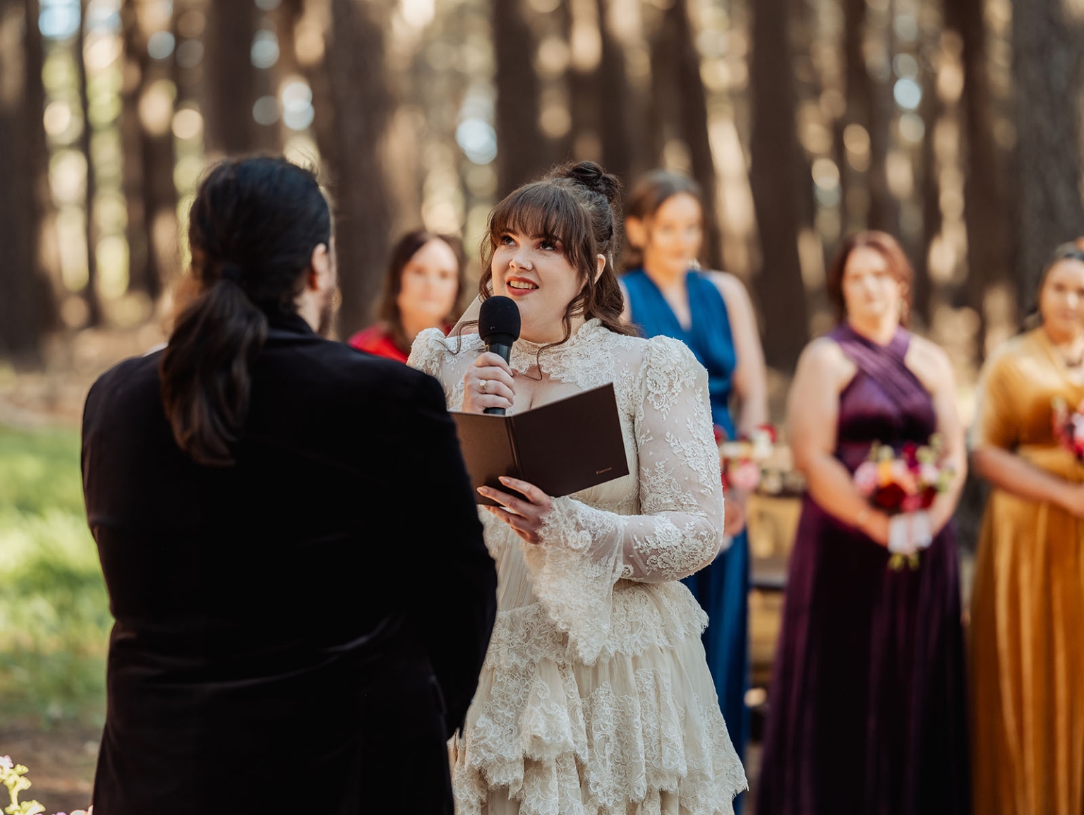 burbrook wedding – amber & angus | kuitpo forest wedding 31 Bride reading her vows into a microphone during a forest wedding ceremony, facing her partner as wedding party members stand behind her.