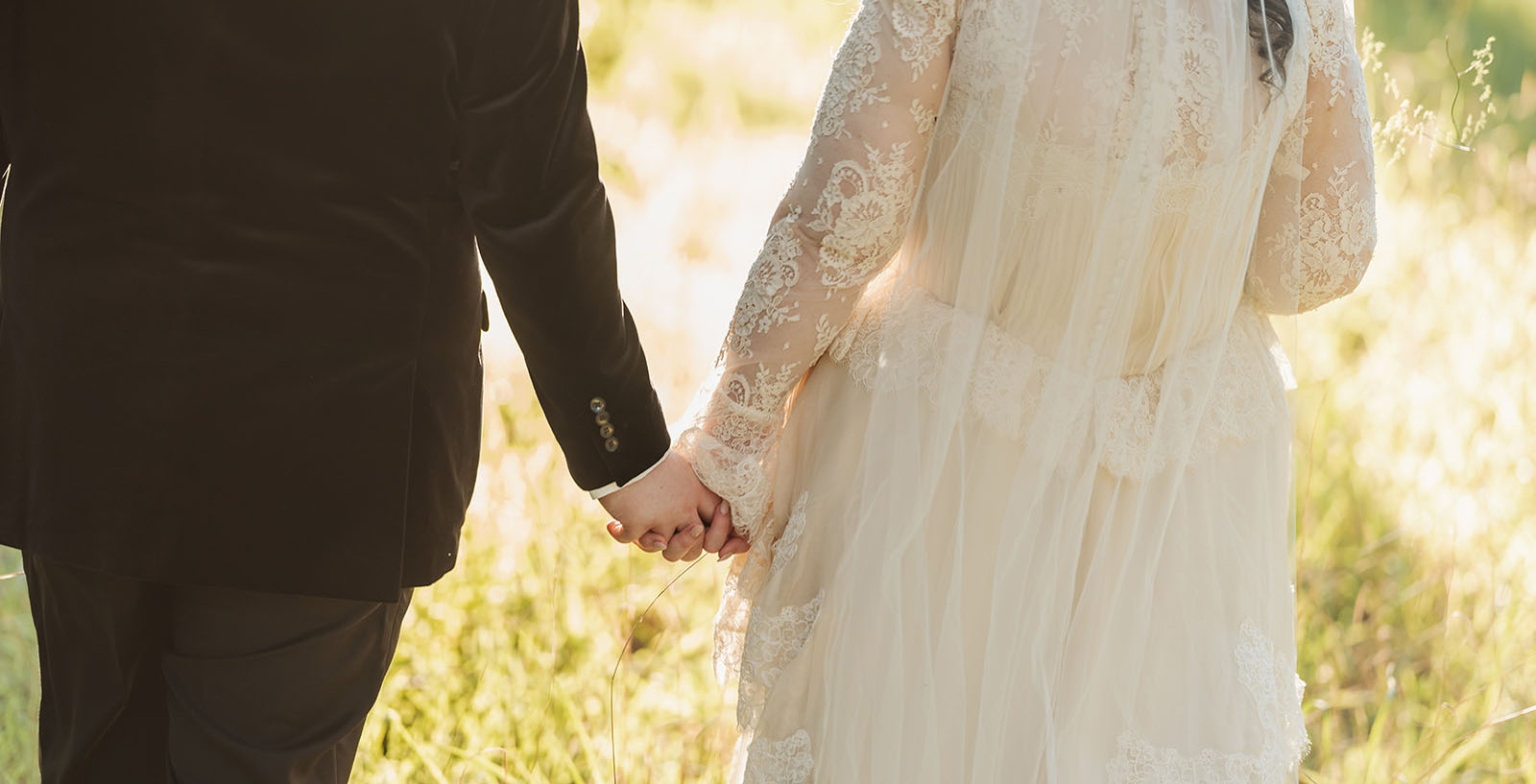 burbrook wedding – amber & angus | kuitpo forest wedding 48 Amber and Angus holding hands, with the bride wearing a lace wedding dress and veil in sunlit grass.