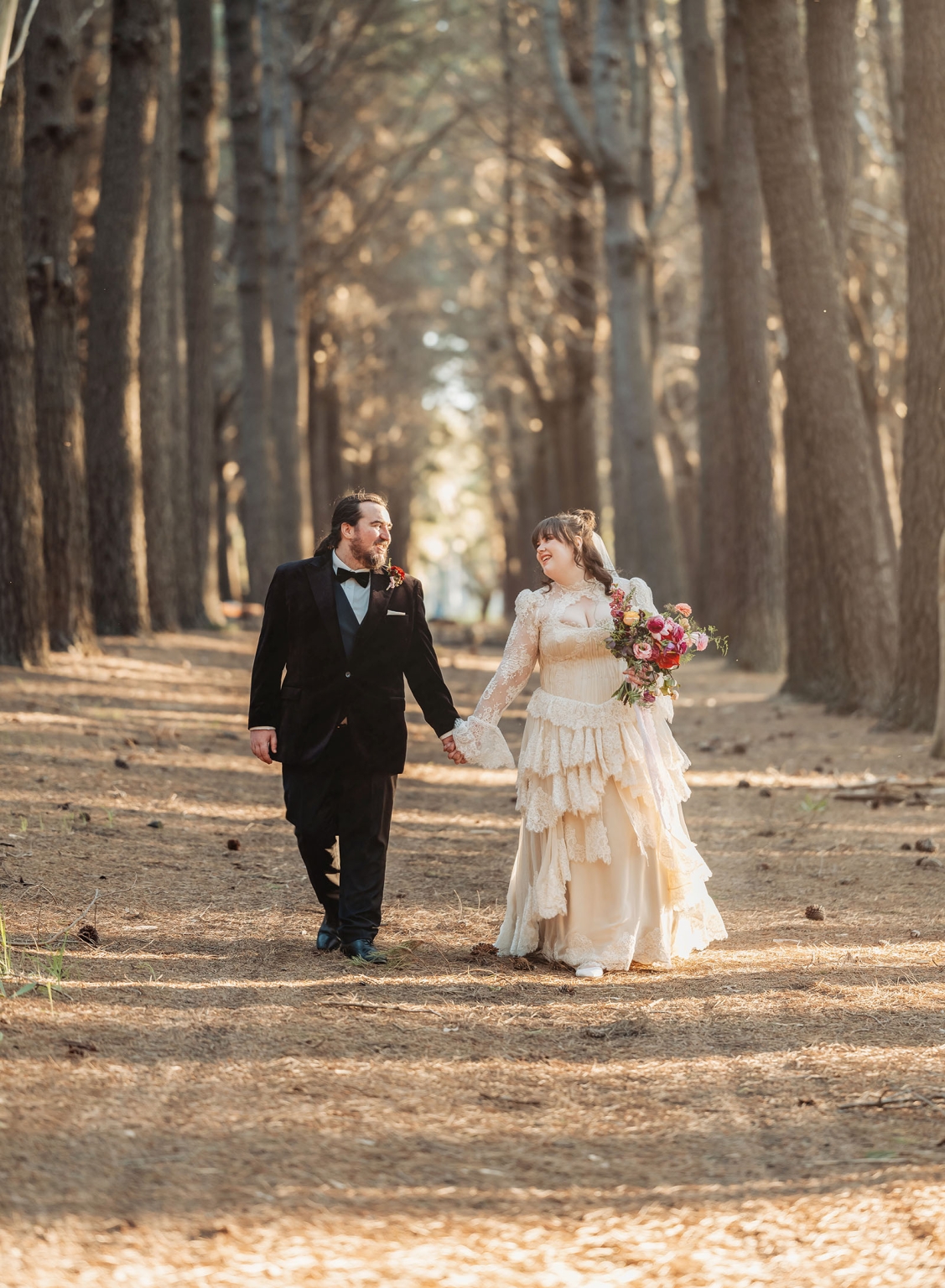 burbrook wedding – amber & angus | kuitpo forest wedding 46 Amber and Angus walk hand in hand beneath tall trees in Kuitpo Forest, with Amber carrying a colourful bouquet.