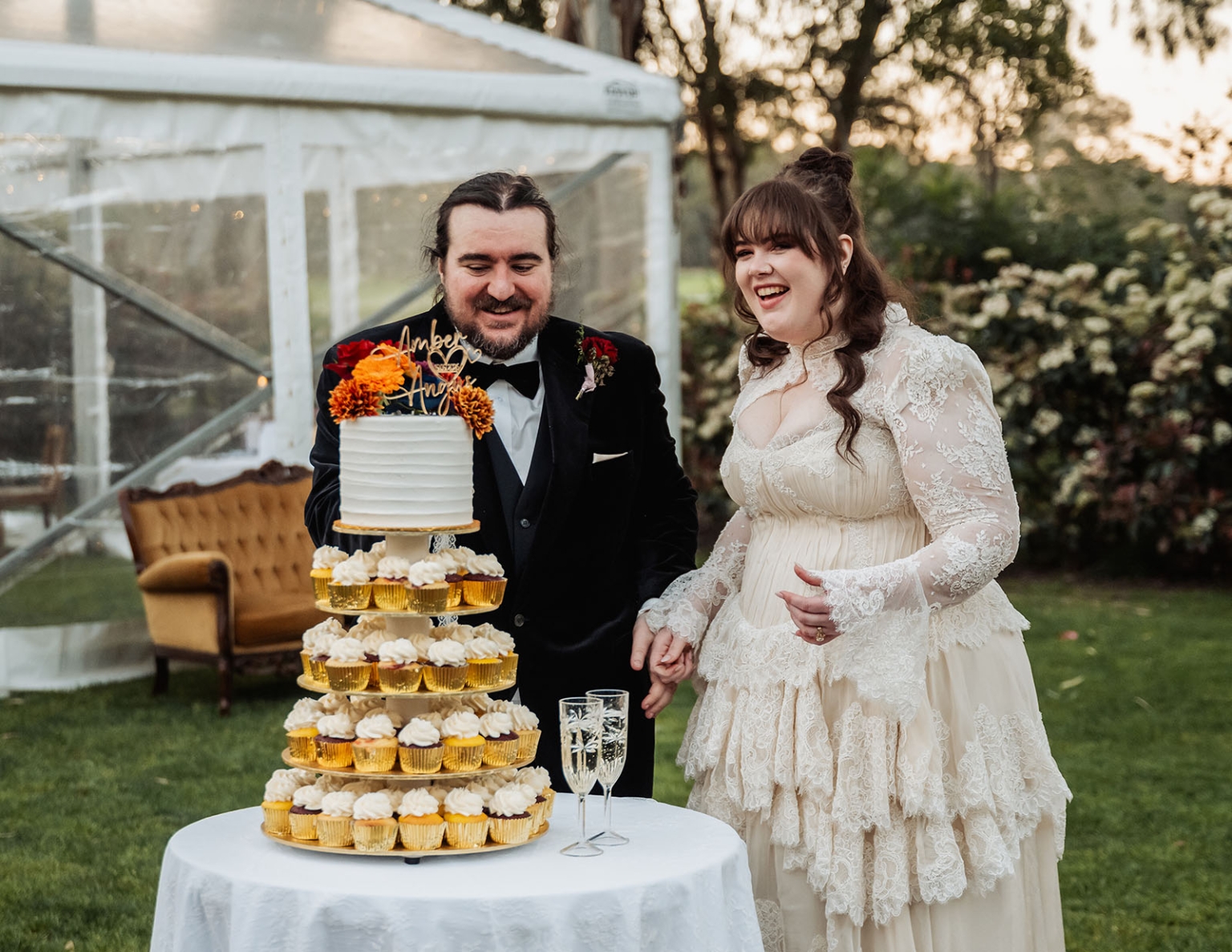 burbrook wedding – amber & angus | kuitpo forest wedding 53 Amber and Angus smiling beside their tiered wedding cake and cupcakes on a lawn at their Kuitpo Forest wedding.