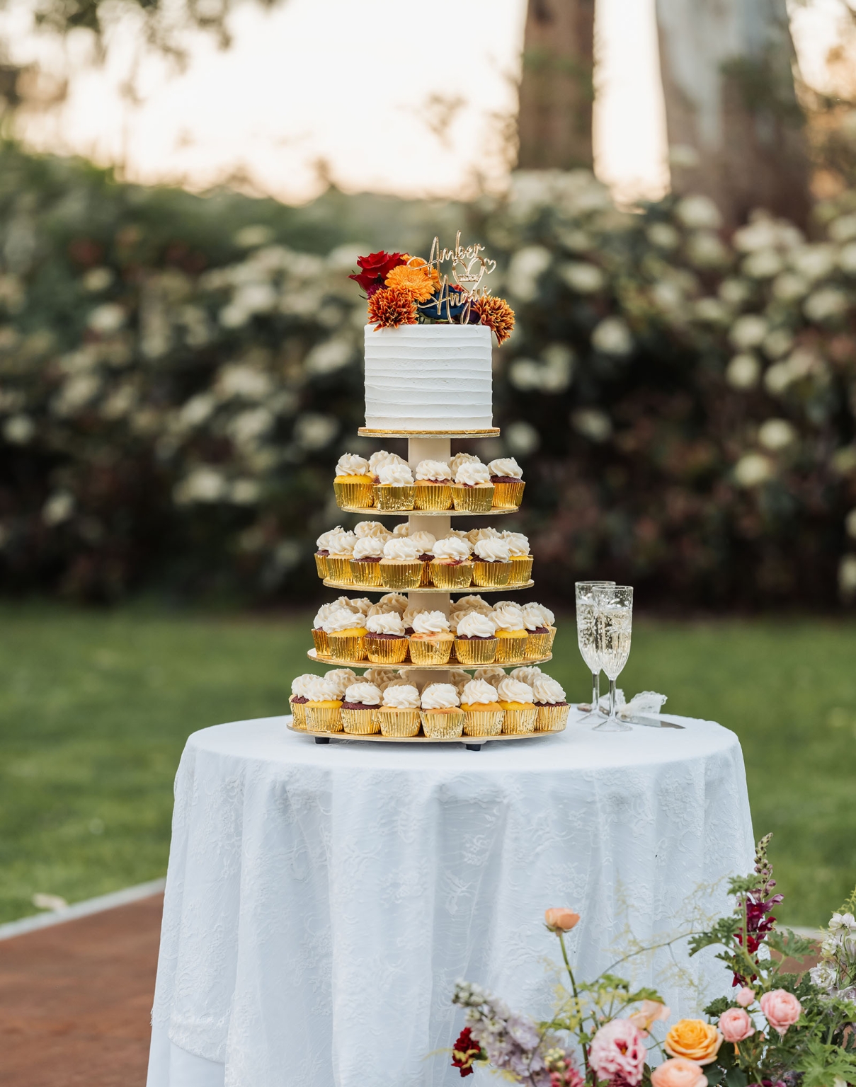 burbrook wedding – amber & angus | kuitpo forest wedding 51 White wedding cake topped with orange and red flowers, displayed above tiers of iced cupcakes on an outdoor table
