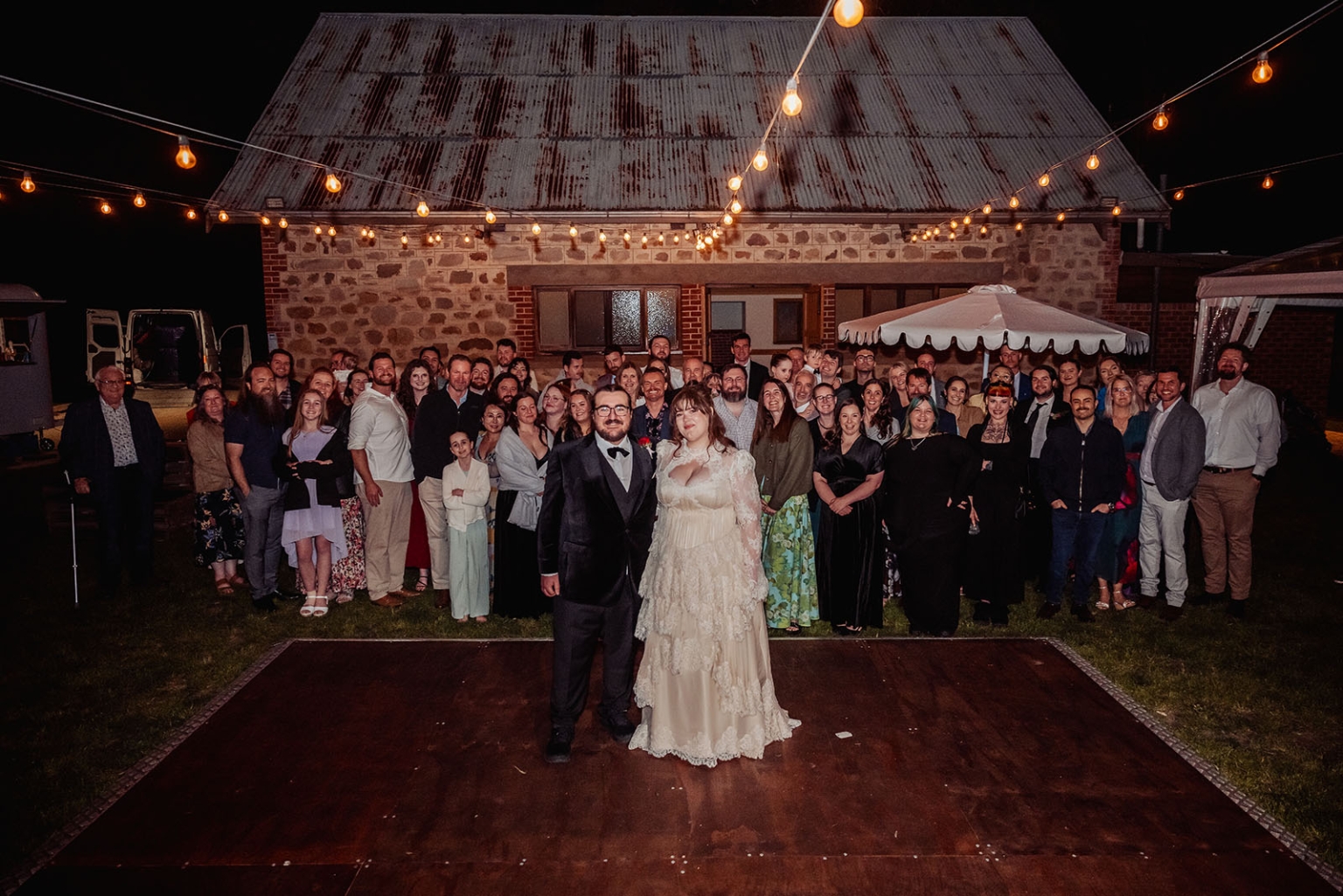burbrook wedding – amber & angus | kuitpo forest wedding 62 Amber and Angus stand with their wedding guests outside a stone hall under festoon lights at night.